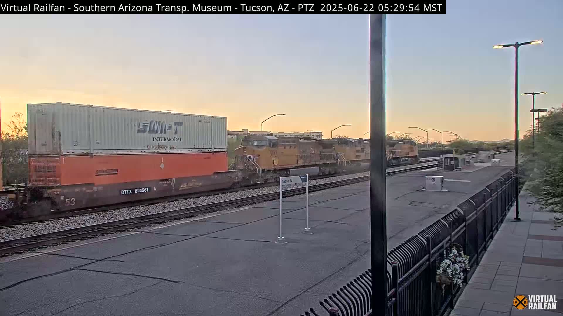 A freight train with a large orange shipping container passes by a train station at sunrise.
