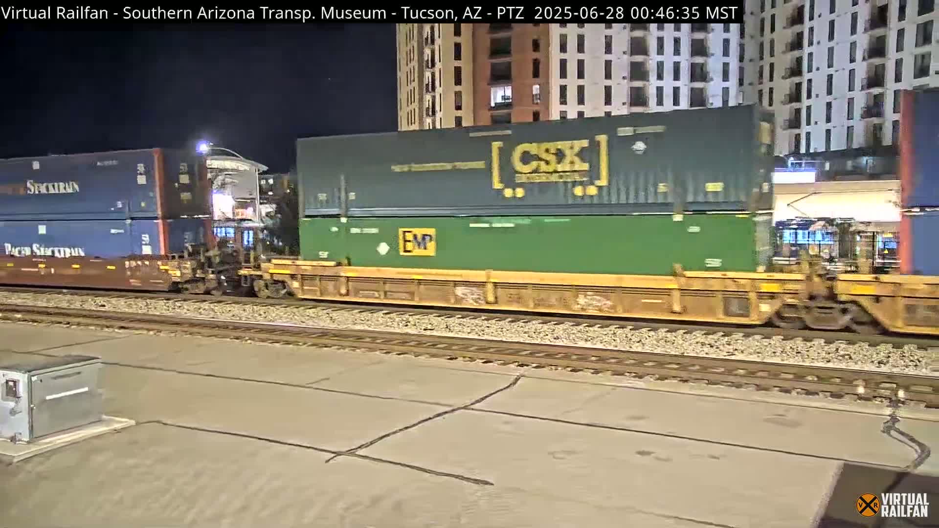 A freight train carrying various shipping containers moves along railroad tracks at night near a multi-story building.