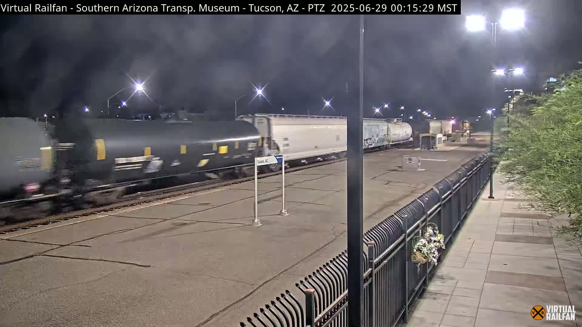 A long freight train passes by a nighttime railway station, under dark, clear skies.
