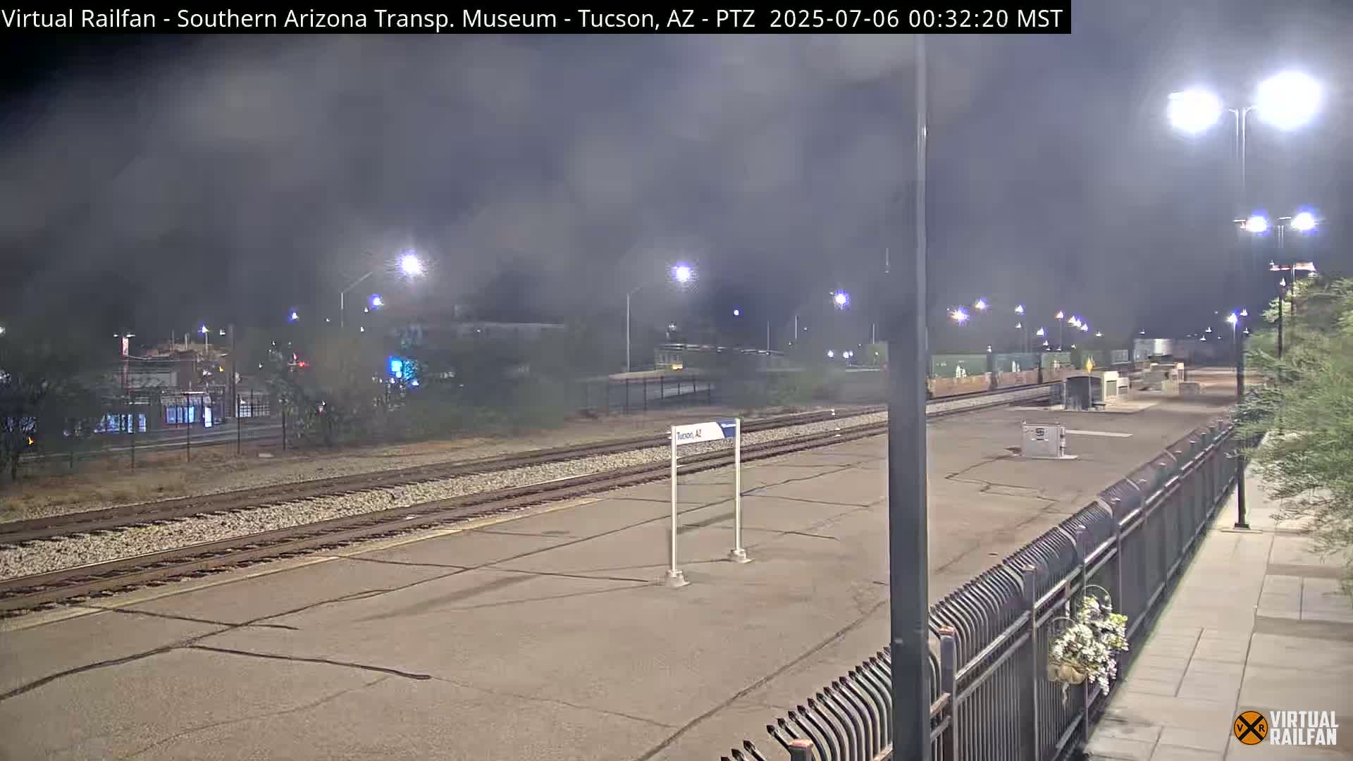 A nighttime view of a mostly empty train station platform under a hazy, dark sky illuminated by streetlights.