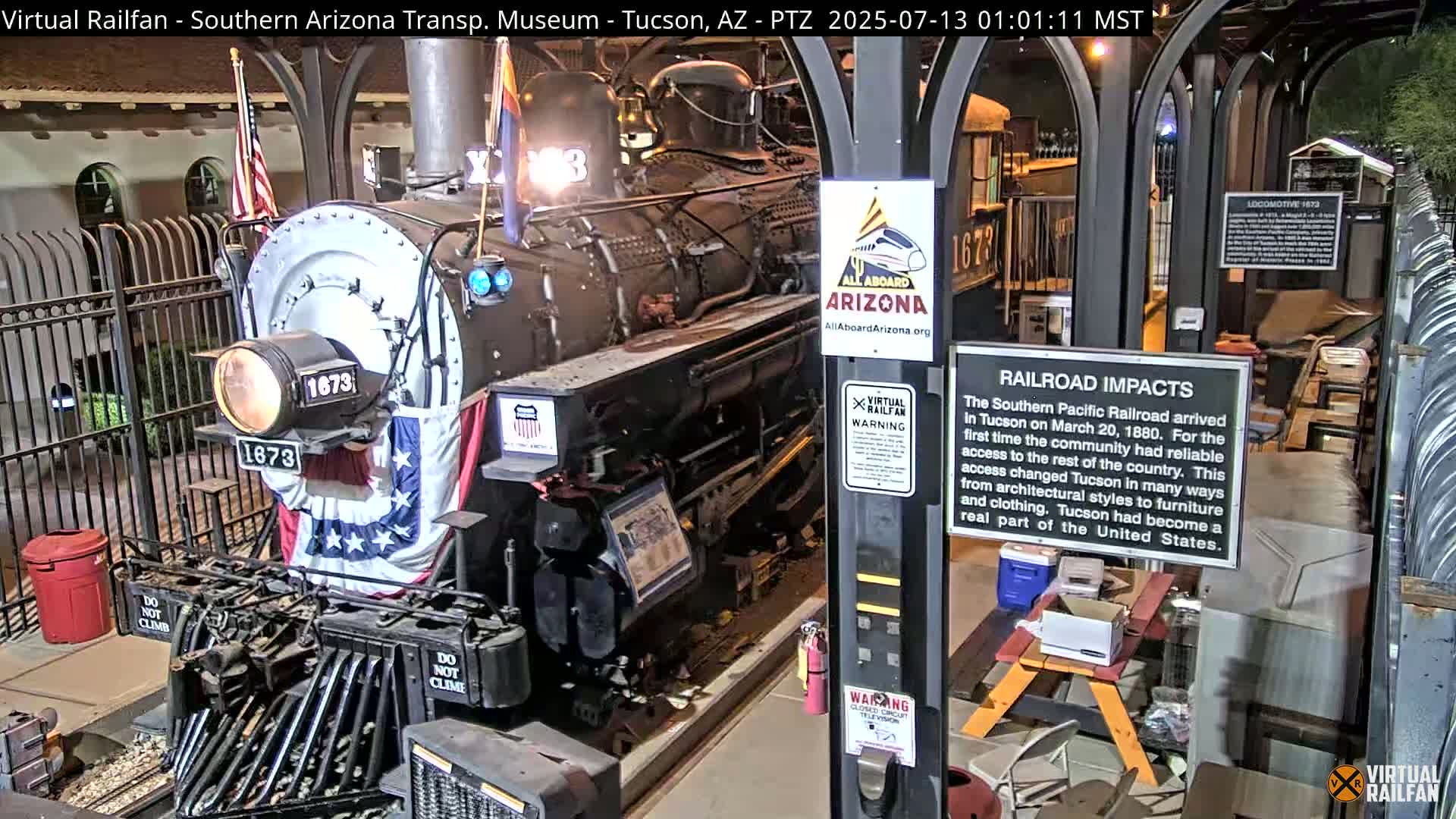 A dark-colored, vintage steam locomotive, adorned with an American flag, sits under nighttime conditions within a sheltered area.