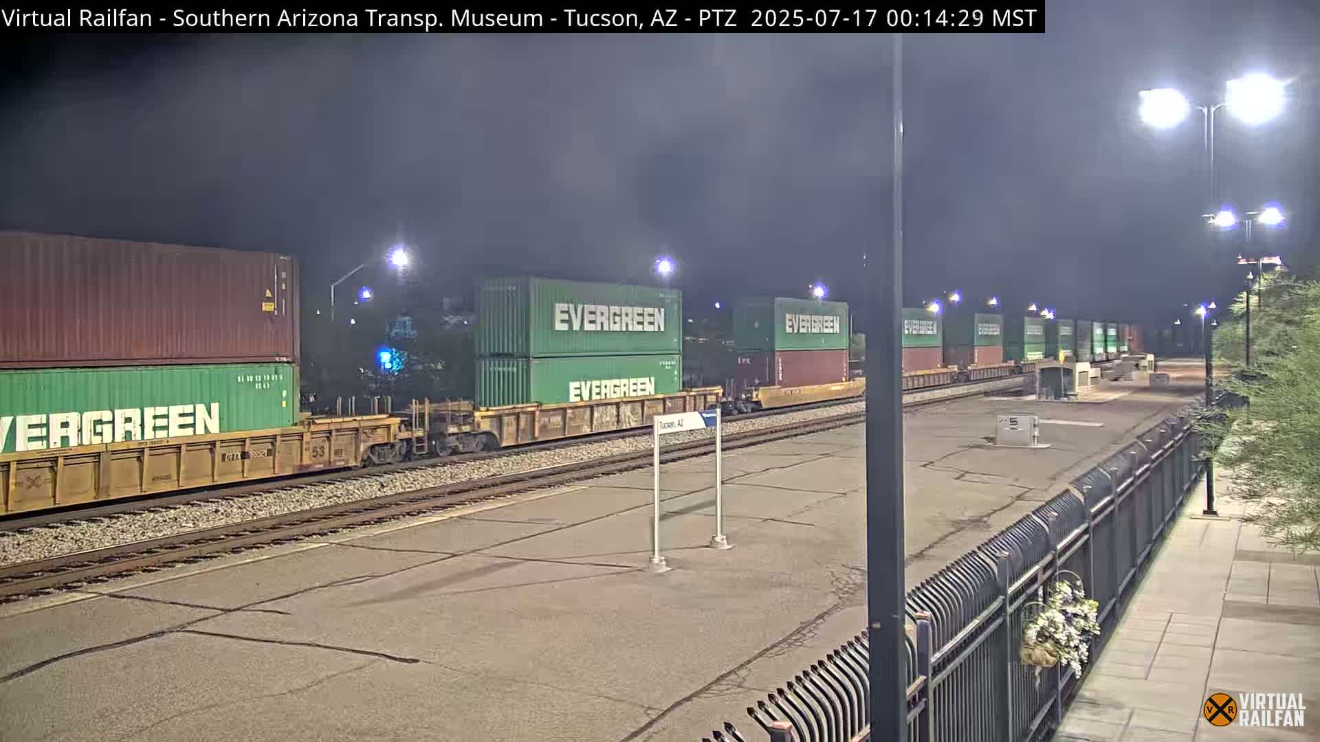 A nighttime view of a train carrying several stacked Evergreen shipping containers sits in a rail yard under bright lights.
