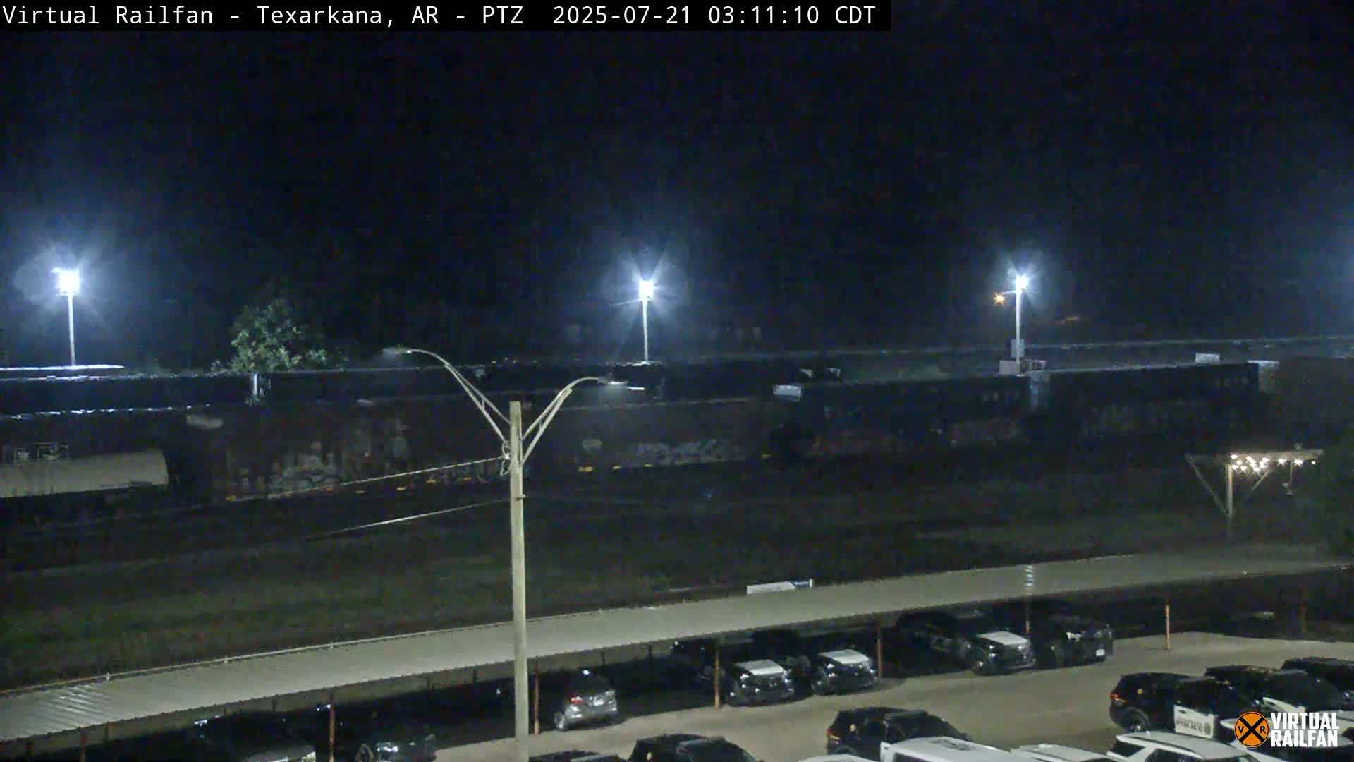 A nighttime view of a train sitting in a rail yard next to a parking lot full of cars under a covered structure.
