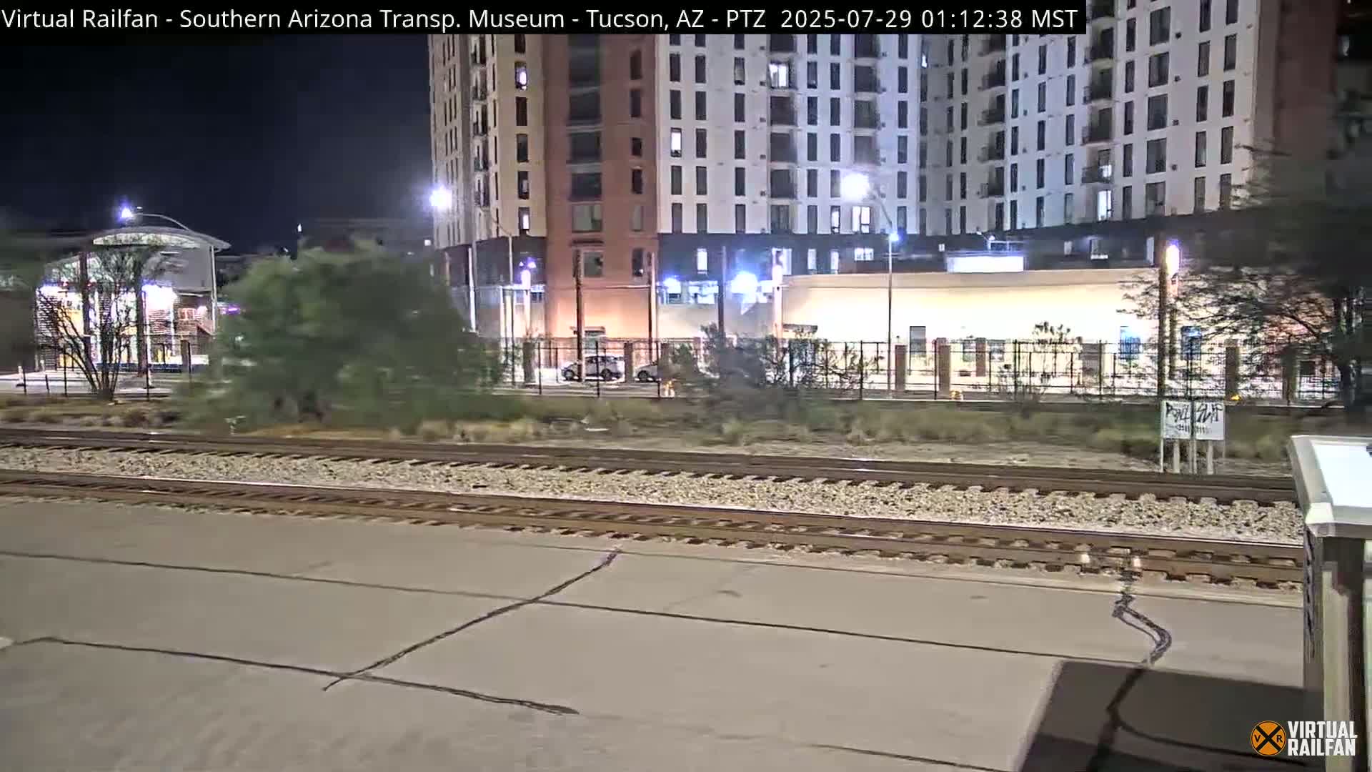 At night, train tracks stretch across a paved area in front of a multi-story building and other structures, under artificial light.