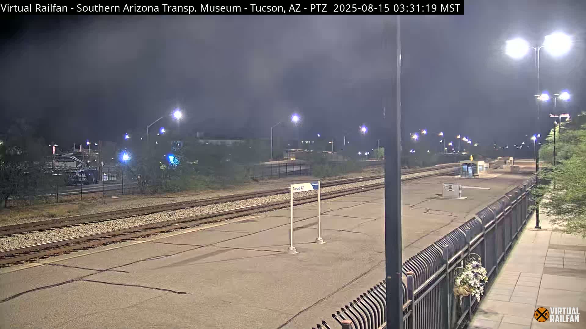 At night, train tracks stretch across a paved area in front of a multi-story building and other structures, under artificial light.