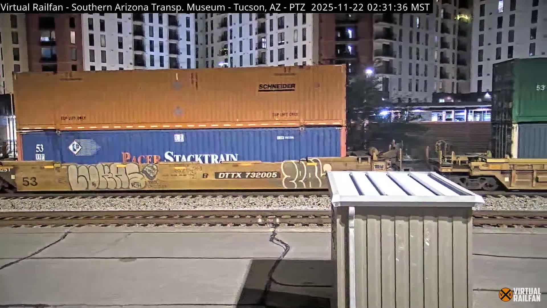 A long freight train carrying double-stacked shipping containers, some featuring prominent graffiti, rests on tracks at night within an urban setting, flanked by illuminated multi-story buildings under clear skies.