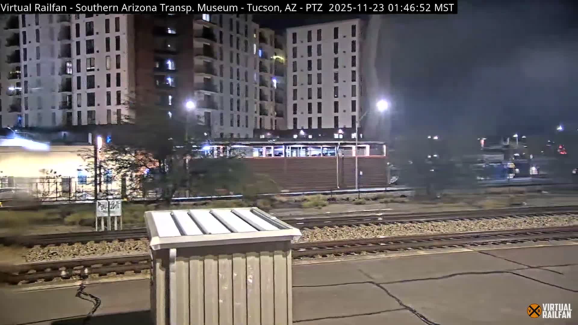 Multiple train tracks are seen in a city scene at night, illuminated by streetlights and building lights, with a station-like structure and tall residential buildings in the background under a clear sky.