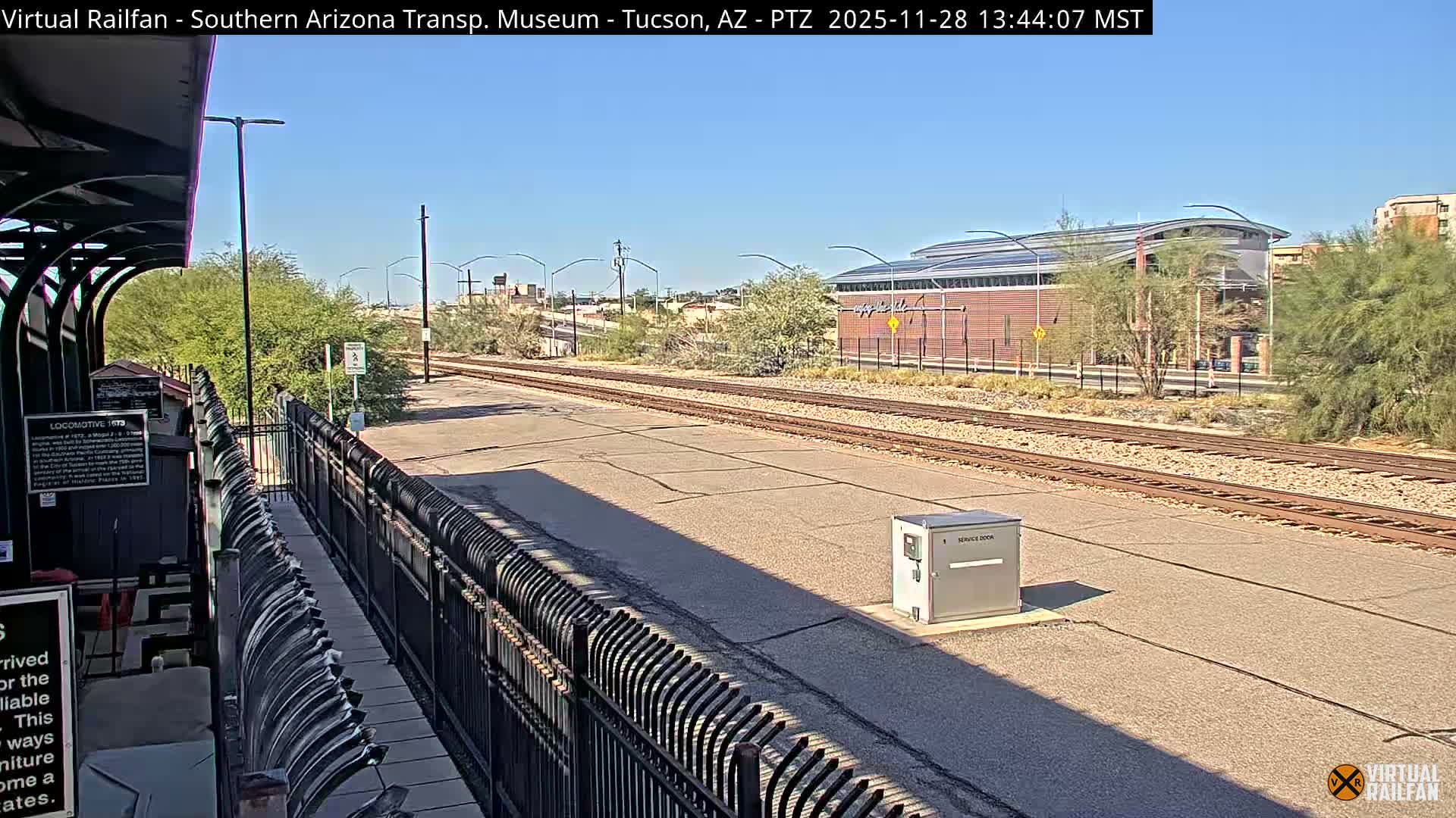A sunny daytime view shows a railroad track beside a paved platform with a black fence, bordered by desert foliage, with a modern building visible in the background under a clear blue sky.
