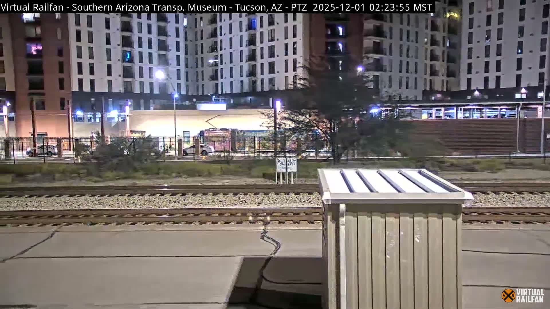 On a clear night, an urban landscape presents train tracks and a utility structure in the foreground, with a street, parked cars, sparse trees, and a graffiti-covered tram in the midground, all set against tall, lit buildings.