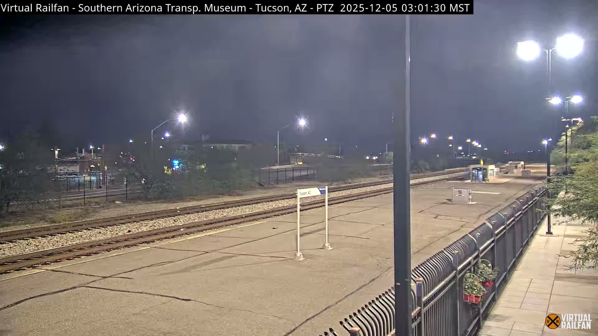 This nighttime image shows a deserted outdoor train platform and adjacent tracks under a clear, calm sky, illuminated by numerous bright streetlights.