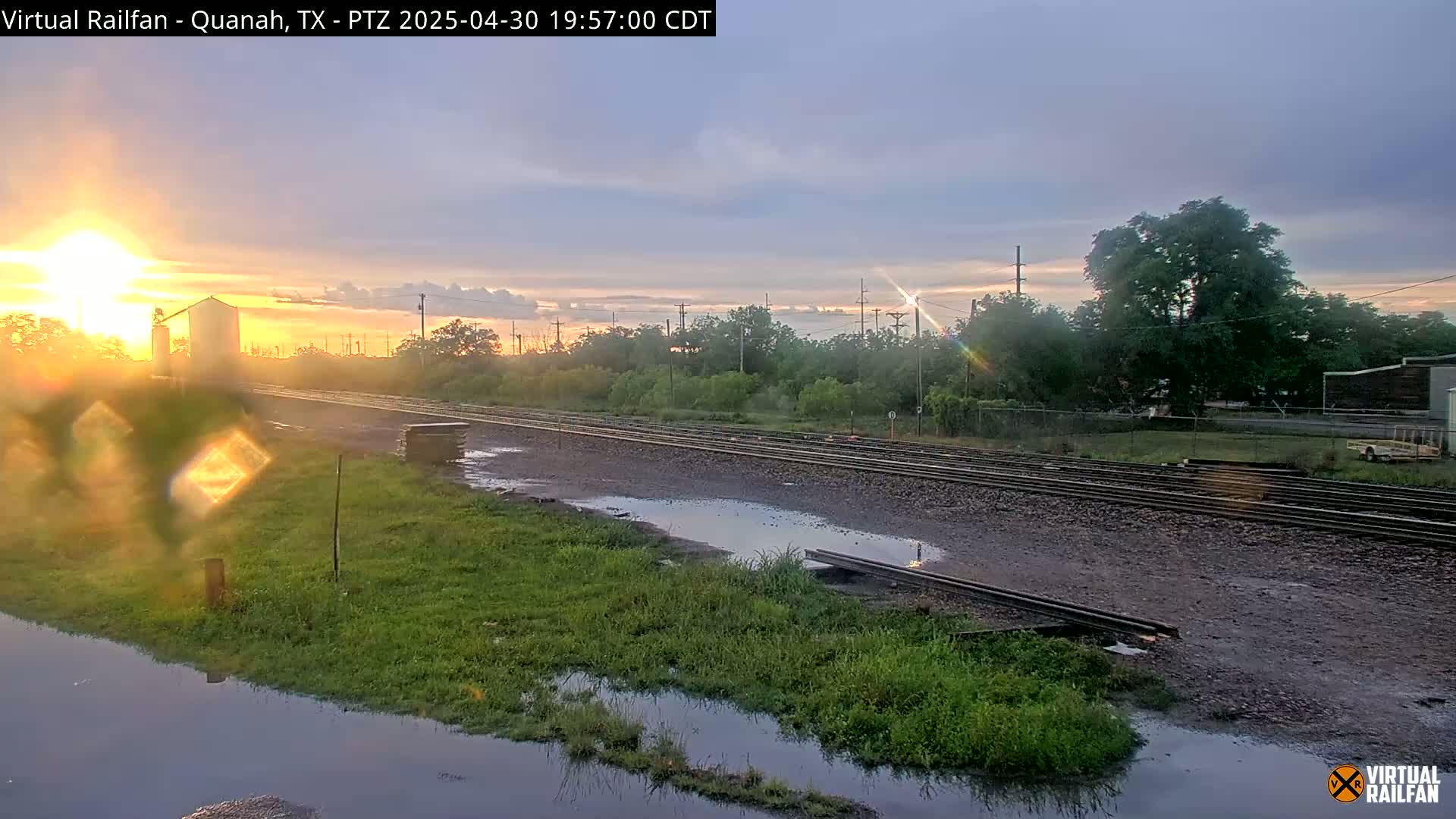 A sunset casts a golden glow over a railway track beside a grassy area with several puddles, near a large building.