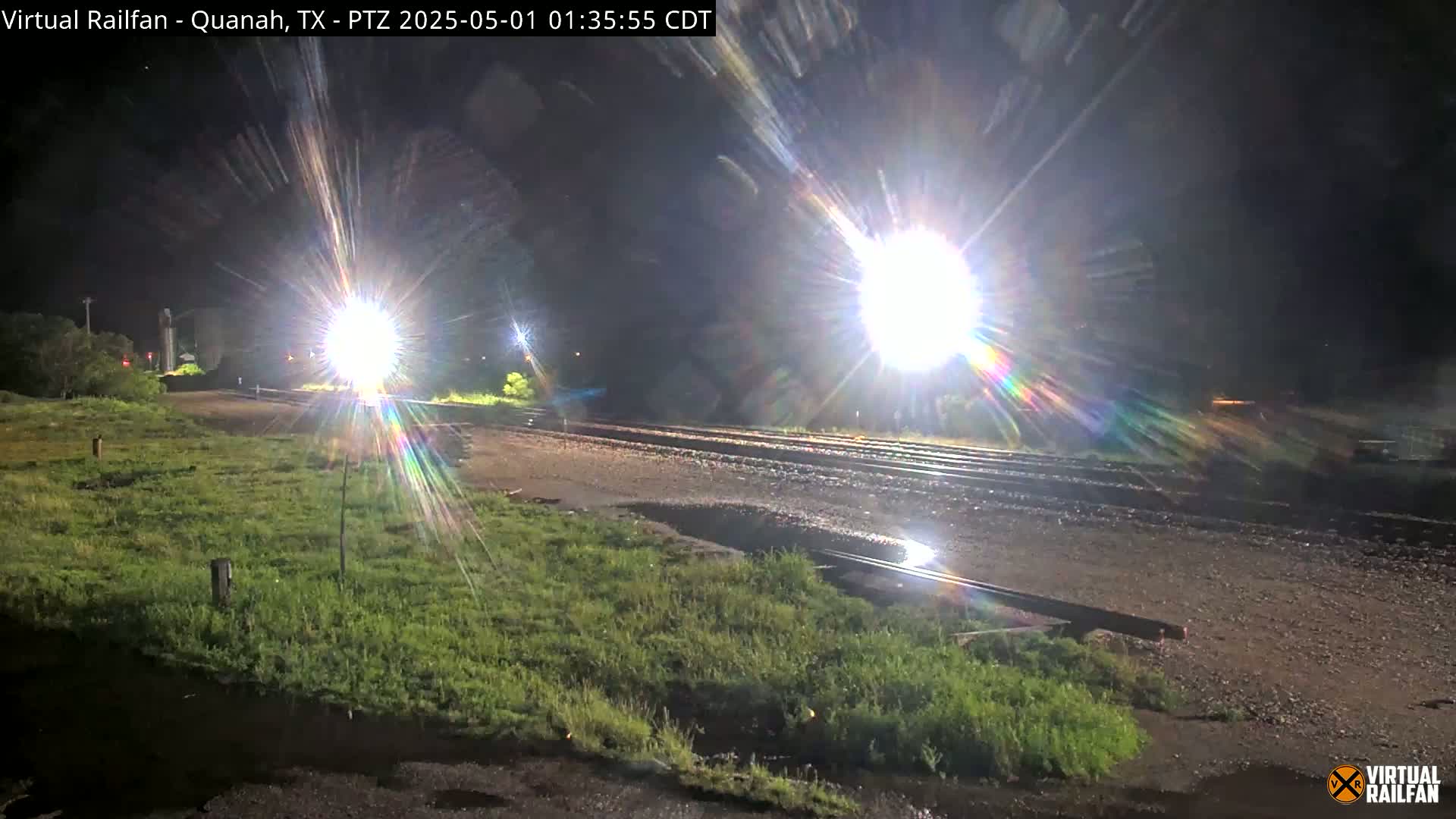 At night, bright lights illuminate a wet, grassy area next to train tracks.