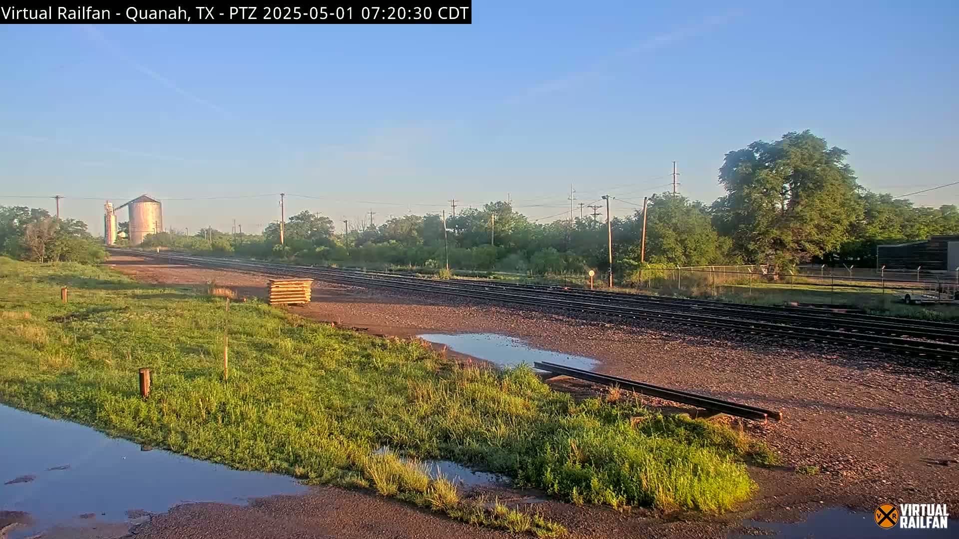 A train track stretches into the distance past a grassy field with puddles, under a clear blue sky, toward a grain silo and trees in the background.