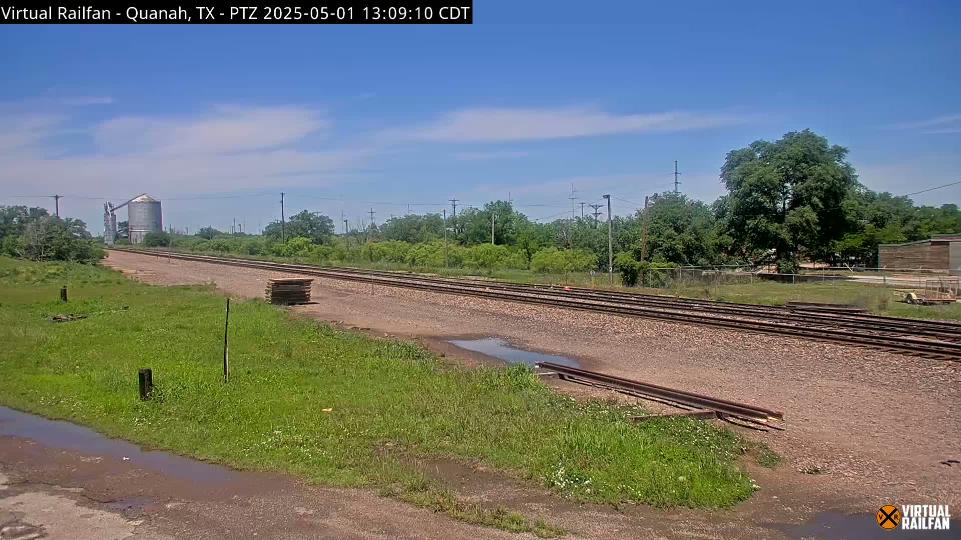 A long stretch of railroad tracks extends into the distance under a mostly clear blue sky, bordered by green vegetation and a few industrial structures.