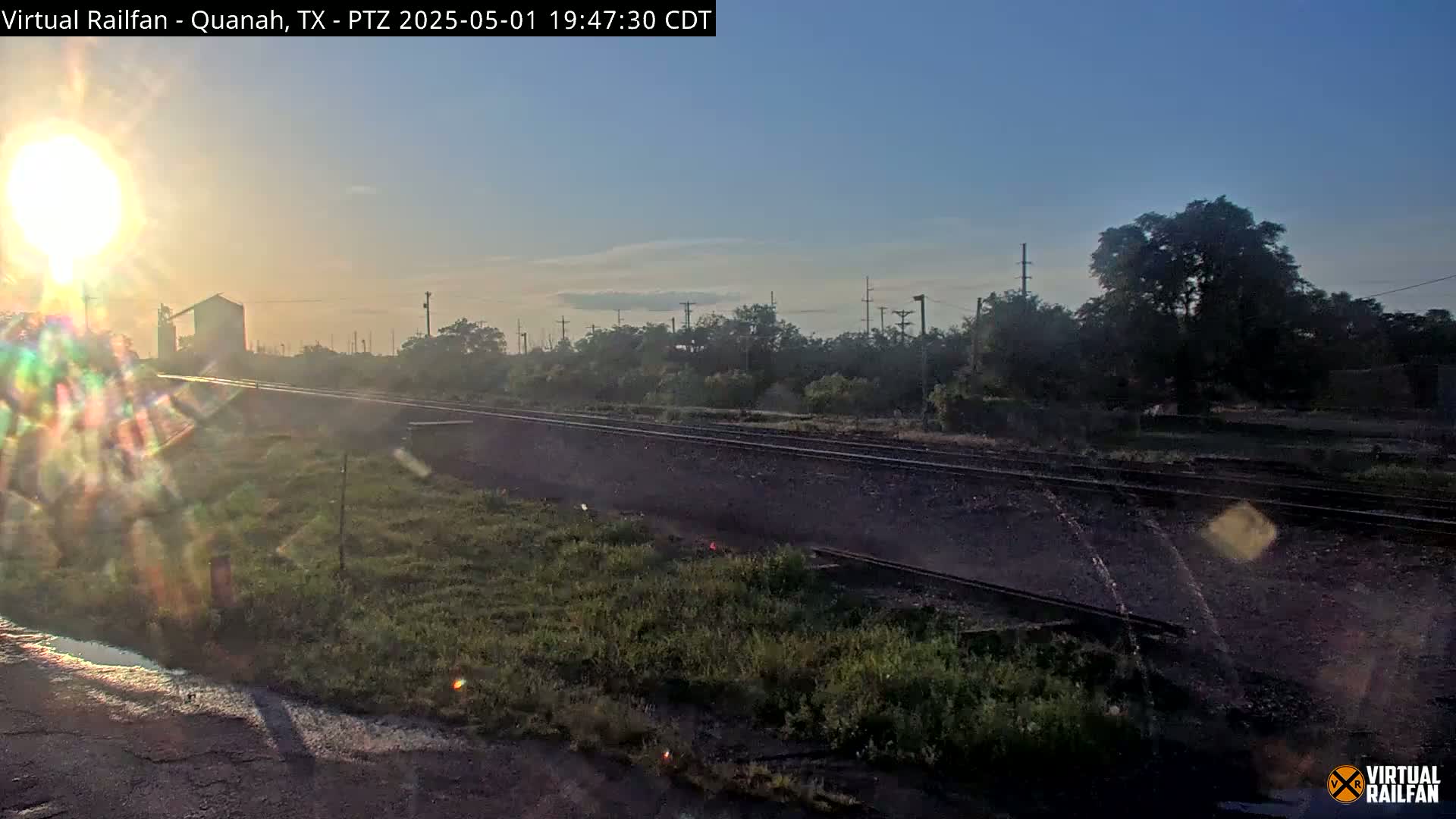 A bright sunrise shines over a railroad track that runs through a grassy area with trees and a silo in the distance.
