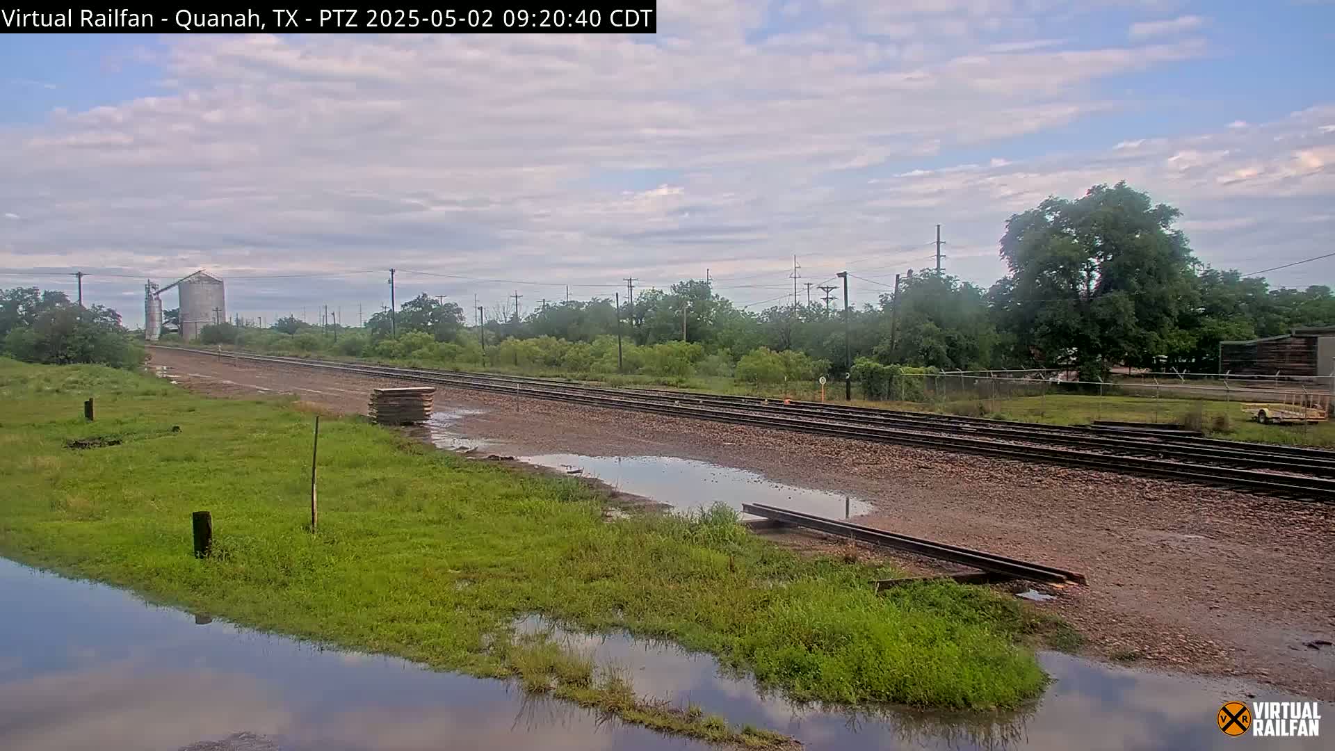 A partly cloudy sky hangs over railroad tracks that run through a grassy area with puddles of water, near a grain silo and some trees.