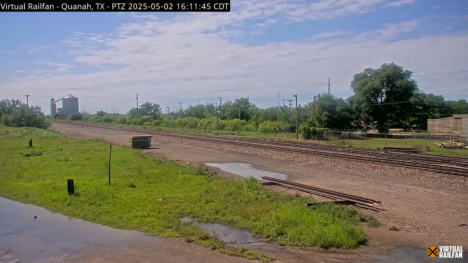 A partly cloudy day reveals railroad tracks extending into the distance, flanked by green grass, a few small puddles, and a grain silo in the far background.