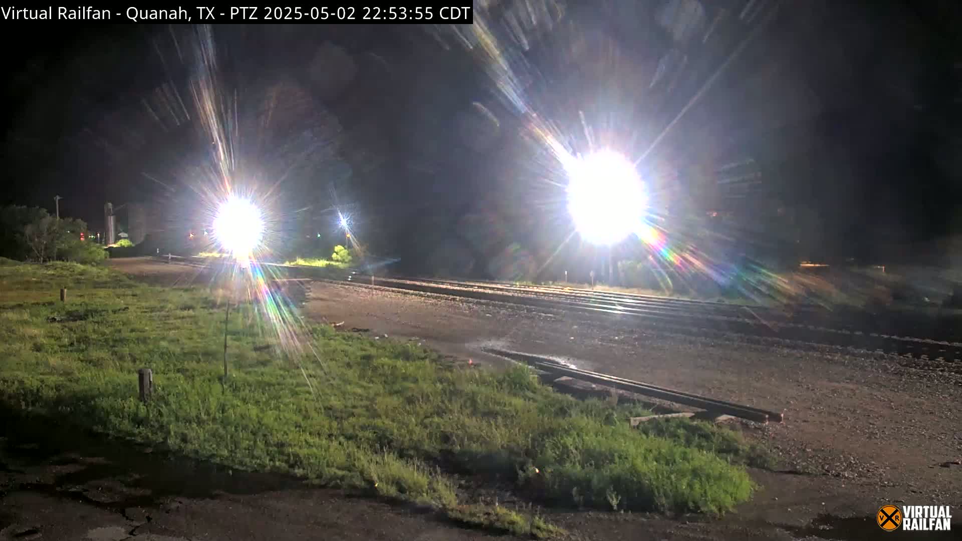Brightly lit train tracks at night are surrounded by short green grass, with several bright lights illuminating the scene.