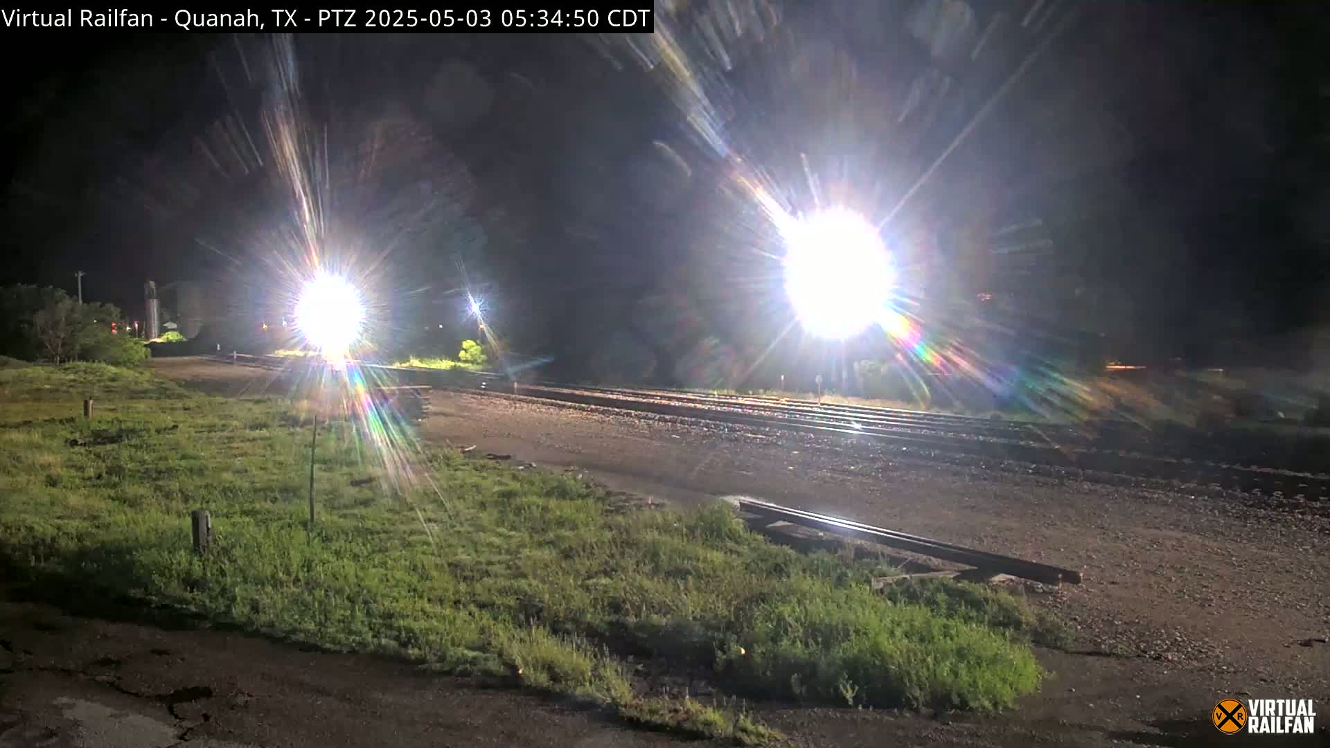 At night, bright lights illuminate a train track bordered by grassy areas.