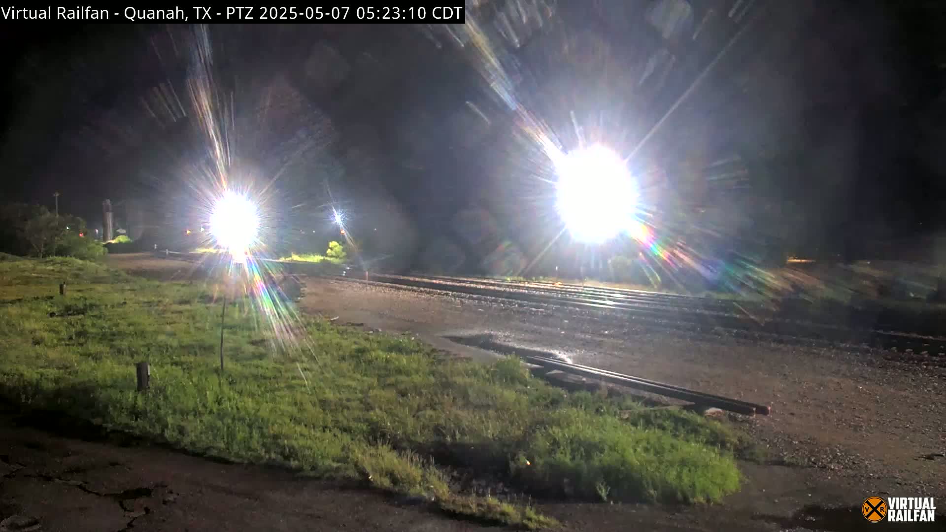 Bright lights illuminate a nighttime scene of railroad tracks and grassy areas, with some wet patches on the ground suggesting recent rain.