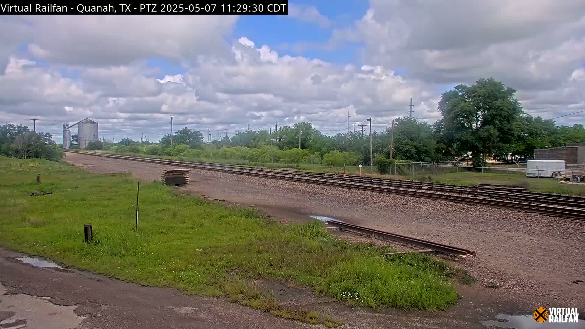 A partly cloudy day shows a railroad track stretching into the distance, passing by green vegetation, a grain silo, and a small building.