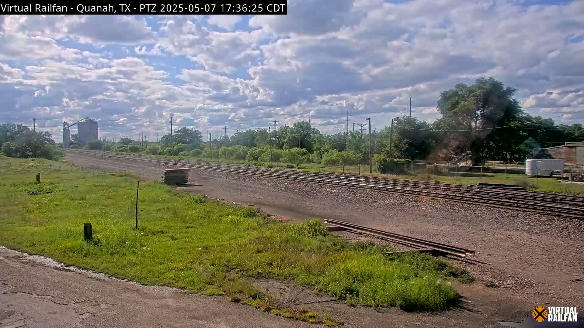 A partly cloudy day shows multiple sets of train tracks stretching into the distance, passing by a grassy area and a grain silo.