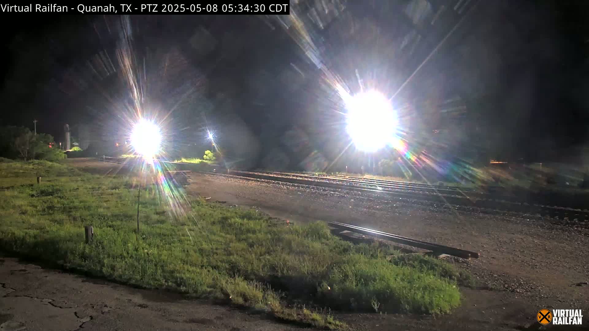 At night, bright lights illuminate a railroad yard with overgrown vegetation surrounding the tracks.