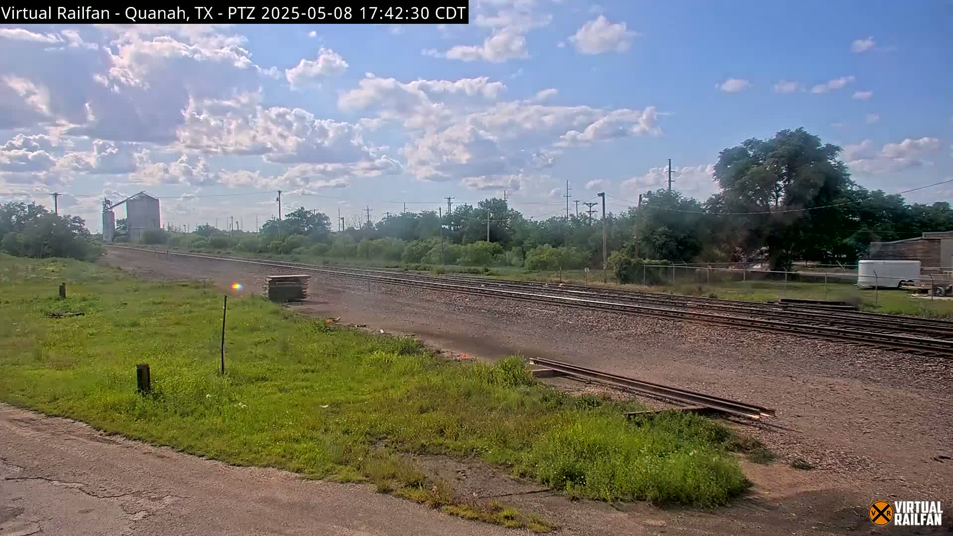 Under a partly cloudy sky, a view of railroad tracks stretches into the distance, passing alongside grassy fields and industrial structures.