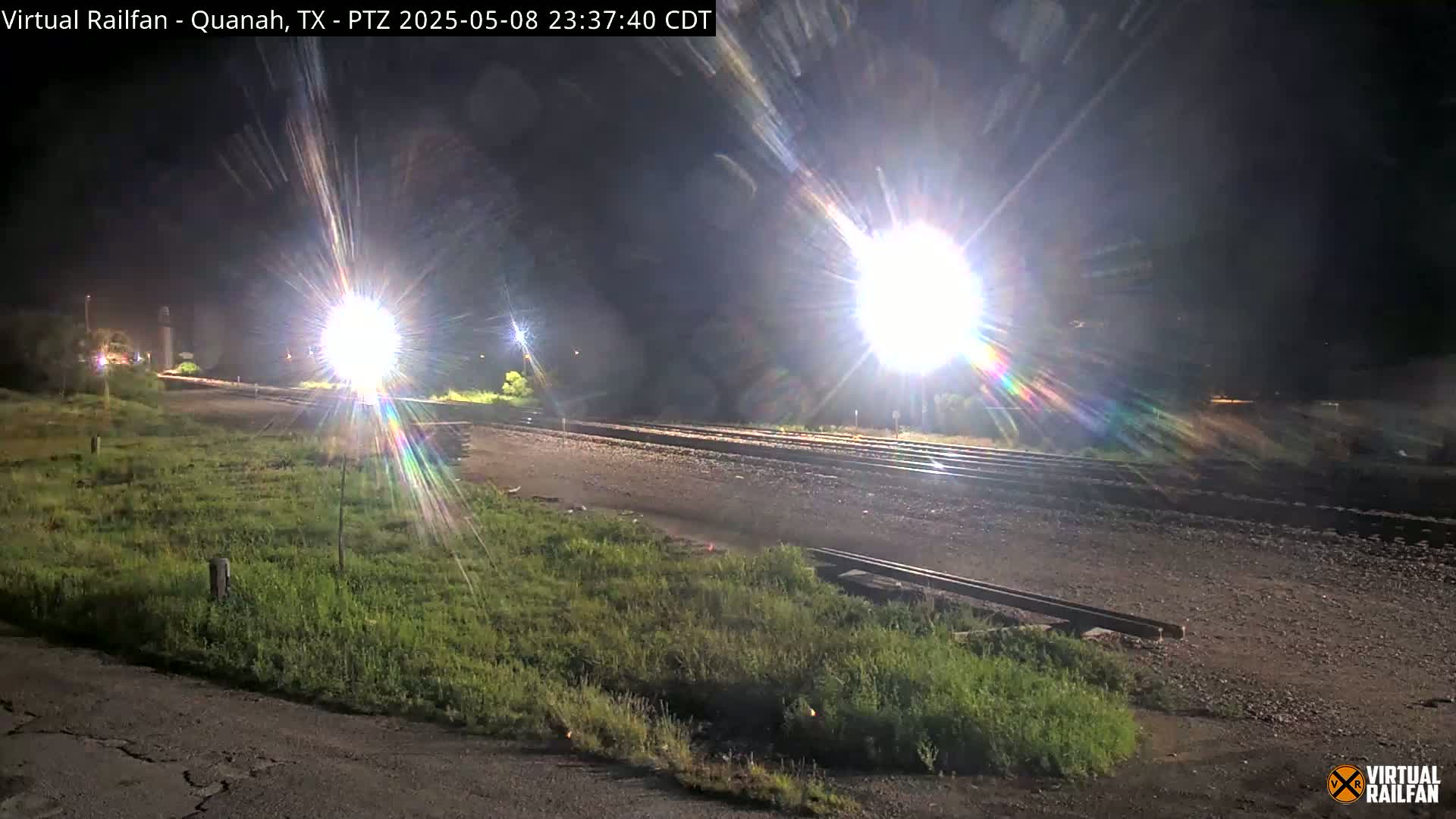 A nighttime view of railroad tracks, brightly lit by powerful lights, bordered by patches of grass.