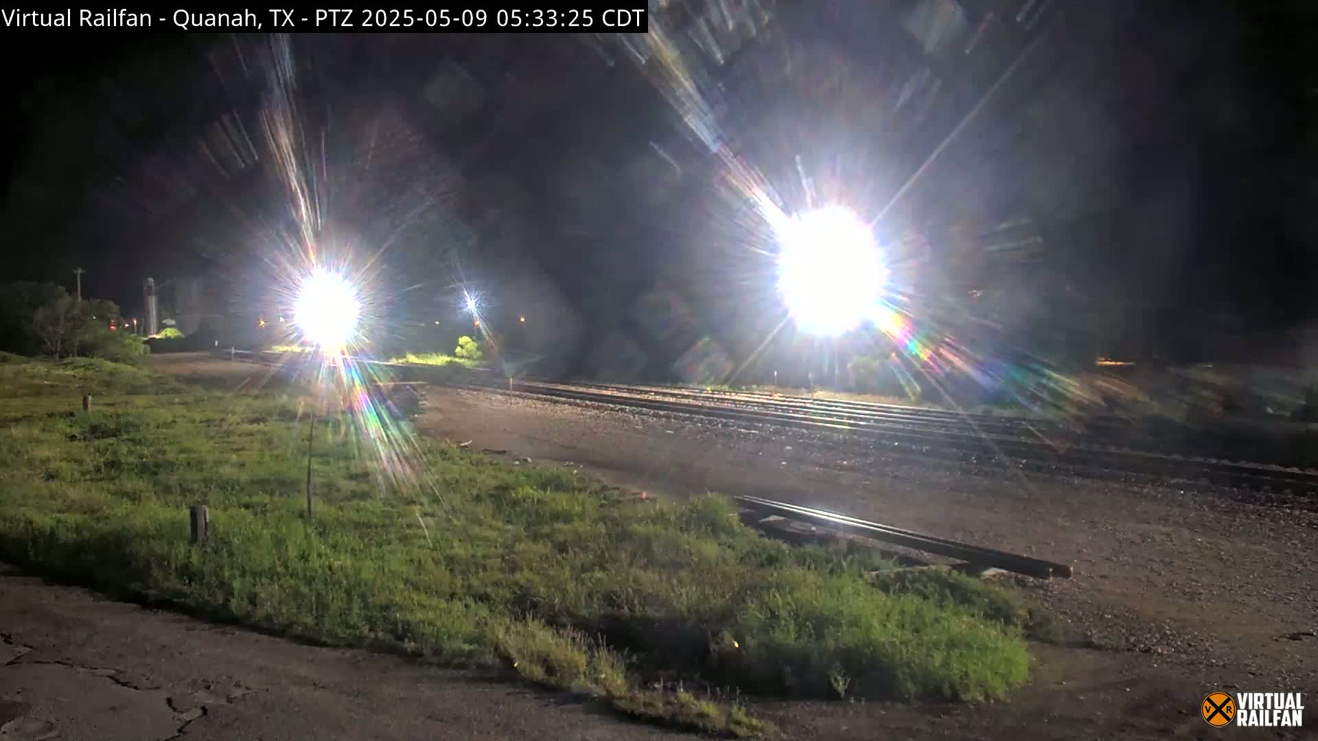 At night, bright lights illuminate a train track surrounded by overgrown grass.