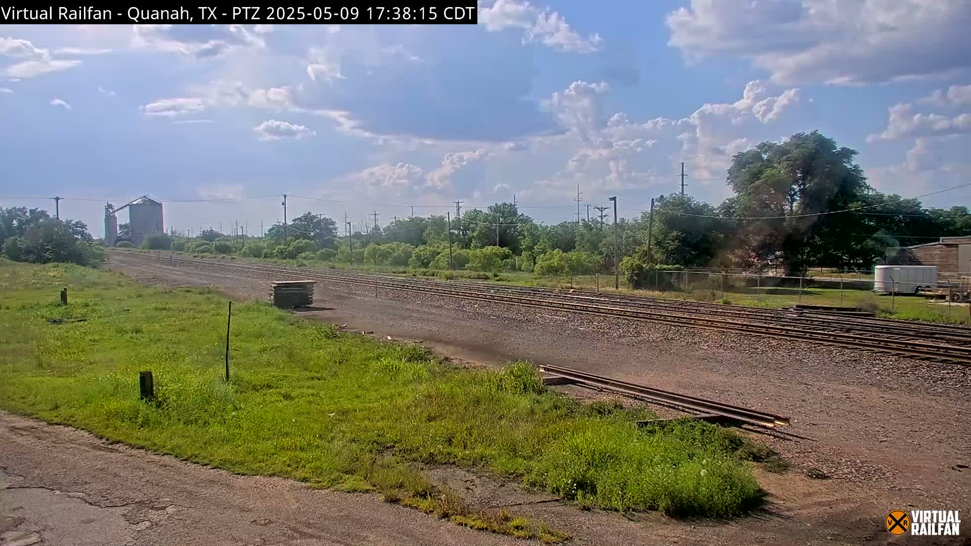A partly cloudy day reveals a view of railroad tracks stretching into the distance, passing beside a grassy area and a grain silo, with trees and other buildings visible in the background.