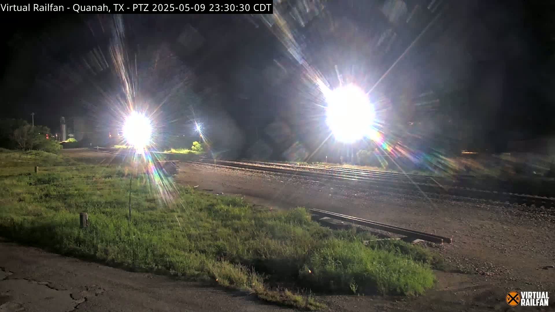 Under a dark night sky, bright lights illuminate a railroad track that runs through grassy areas.