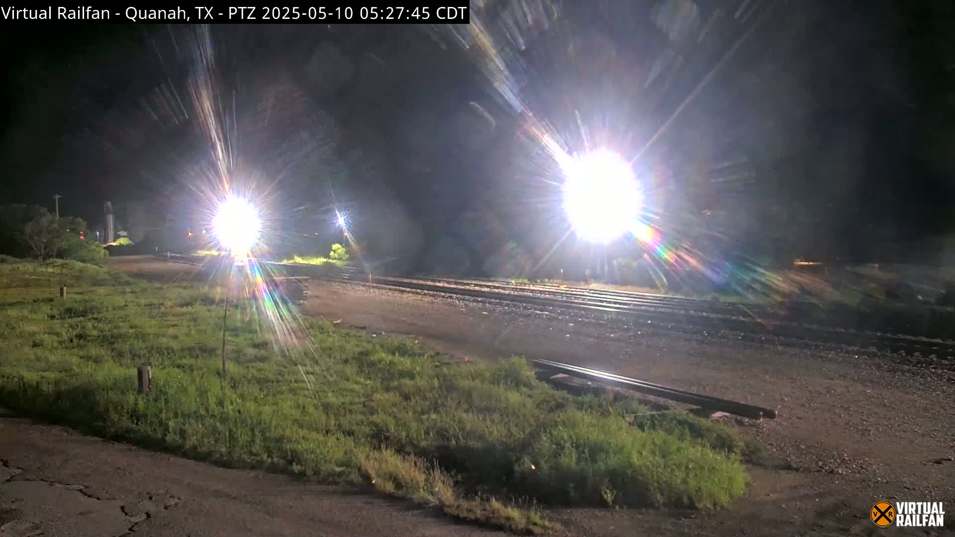 A nighttime view of train tracks illuminated by bright lights, with grassy areas on either side.