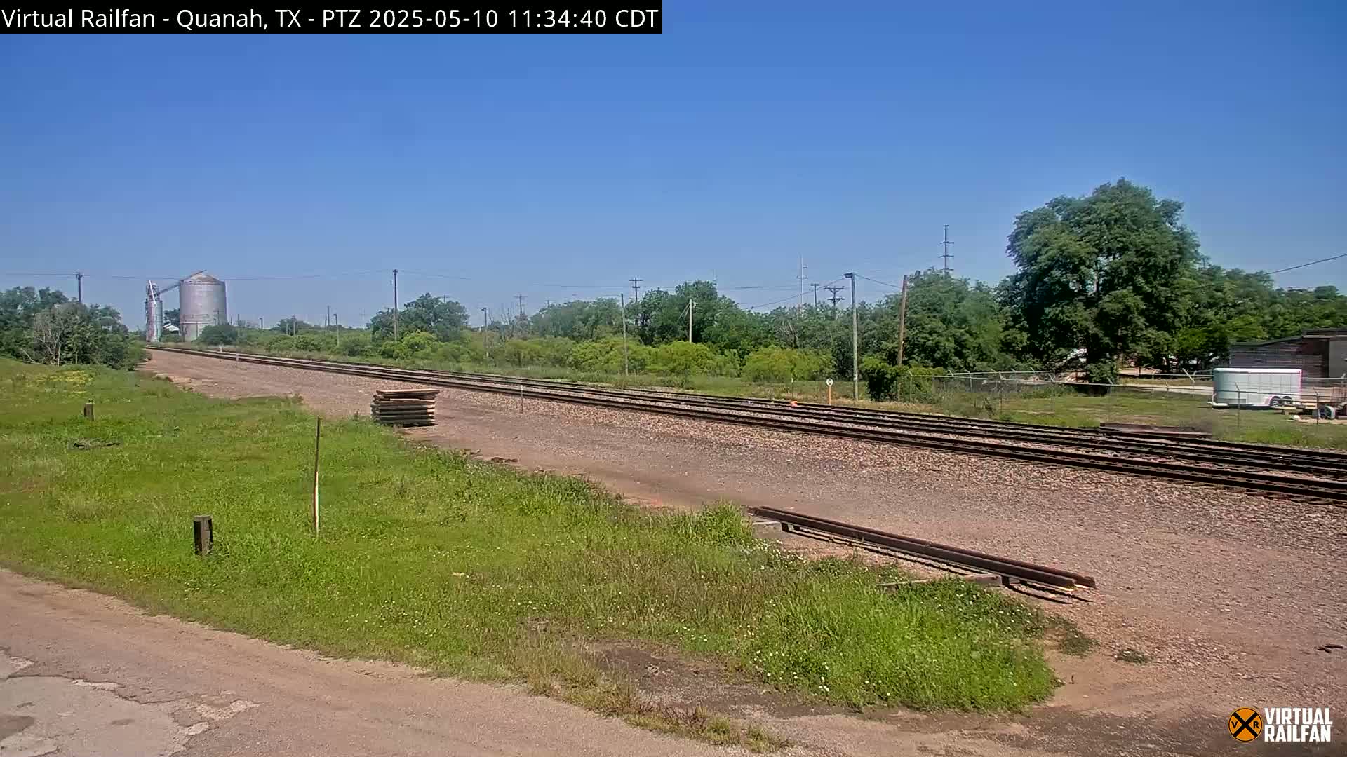 Under a clear blue sky, a stretch of railroad tracks extends into the distance, passing by grassy fields, trees, and a grain silo.