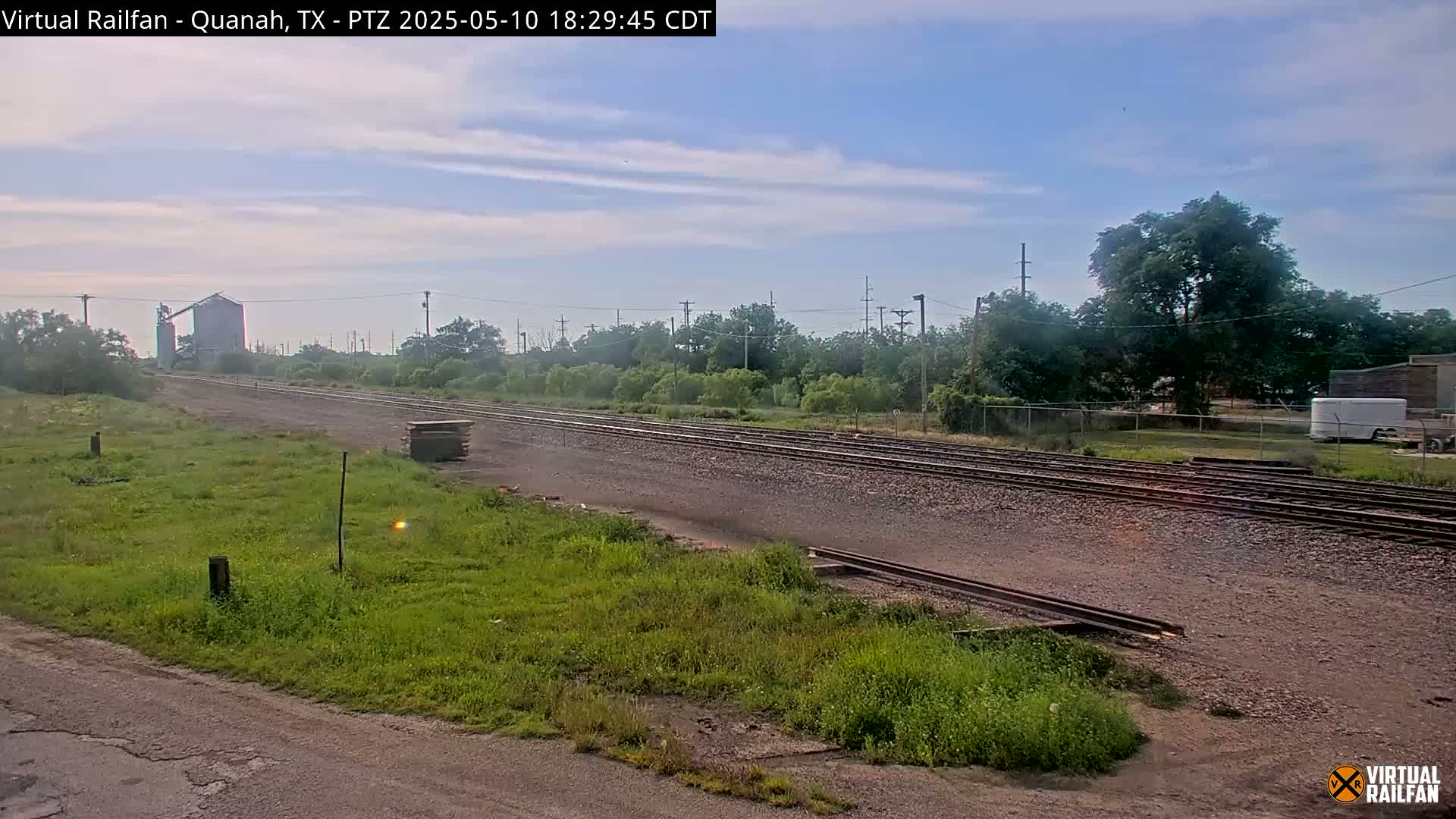 Under a mostly clear sky, a wide shot shows several parallel train tracks stretching into the distance, flanked by grassy areas, trees, and a grain silo in the background.