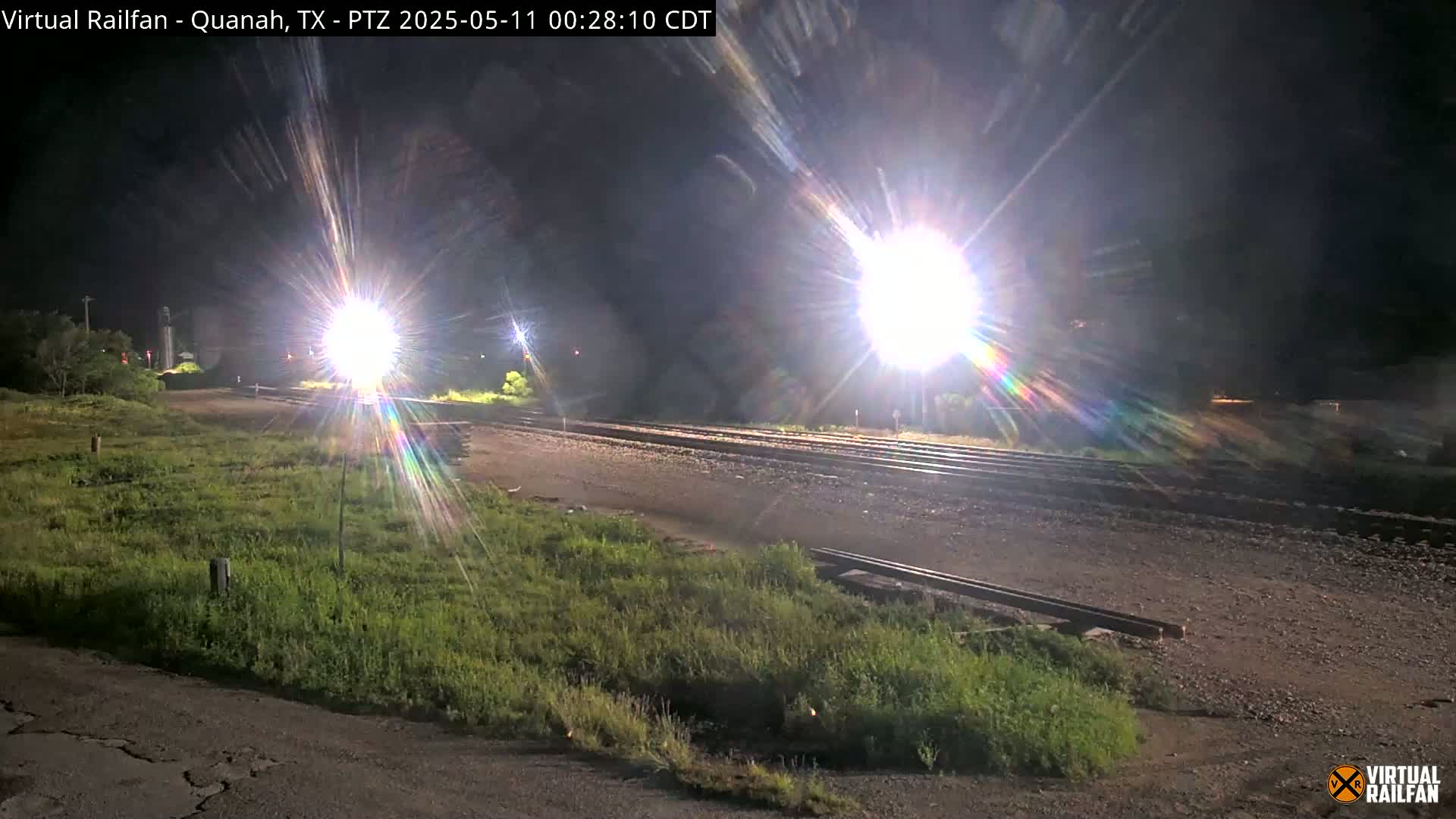 At night, bright lights illuminate a railroad crossing next to grassy areas.