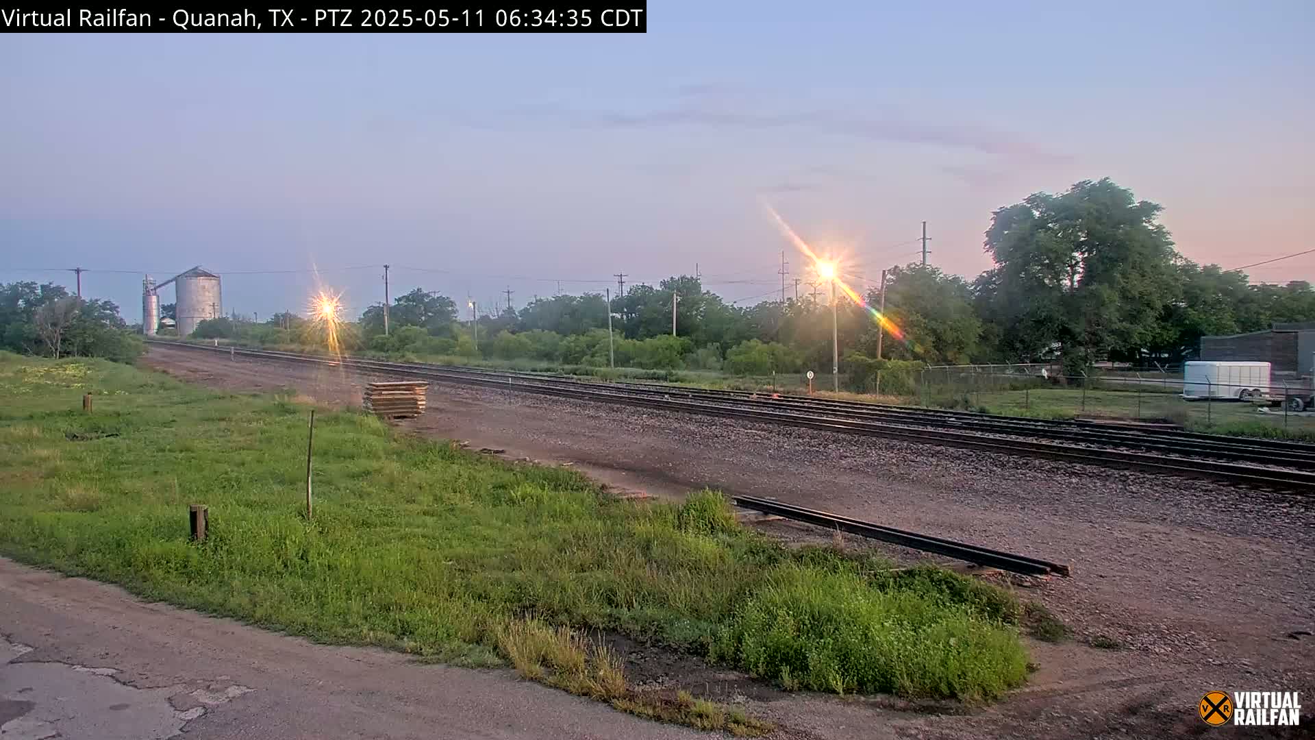 A long stretch of train tracks extends into the distance under a pale sunrise, passing alongside a grassy field and a few trees.