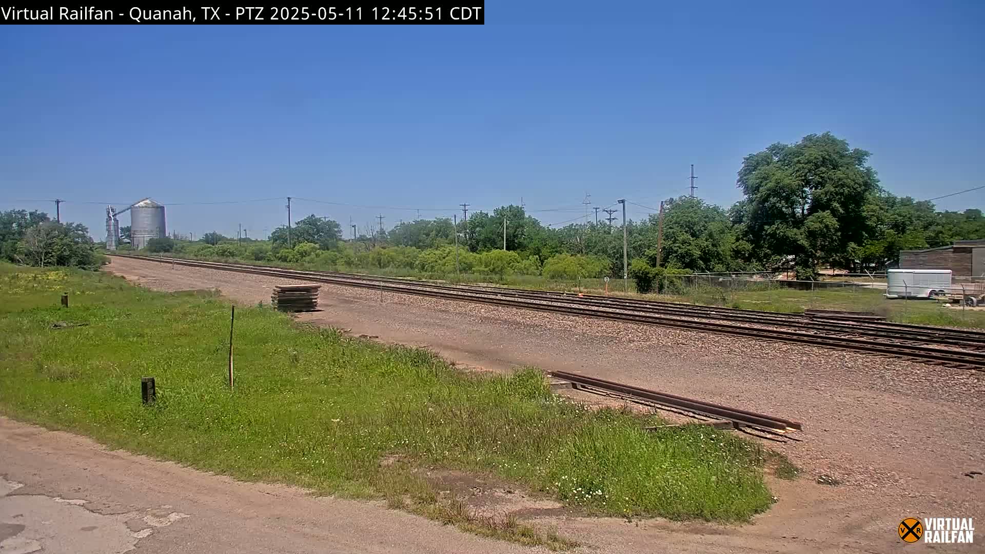A long stretch of train tracks extends into the distance under a clear blue sky, bordered by grassy fields and a line of trees, with a grain silo visible in the far background.