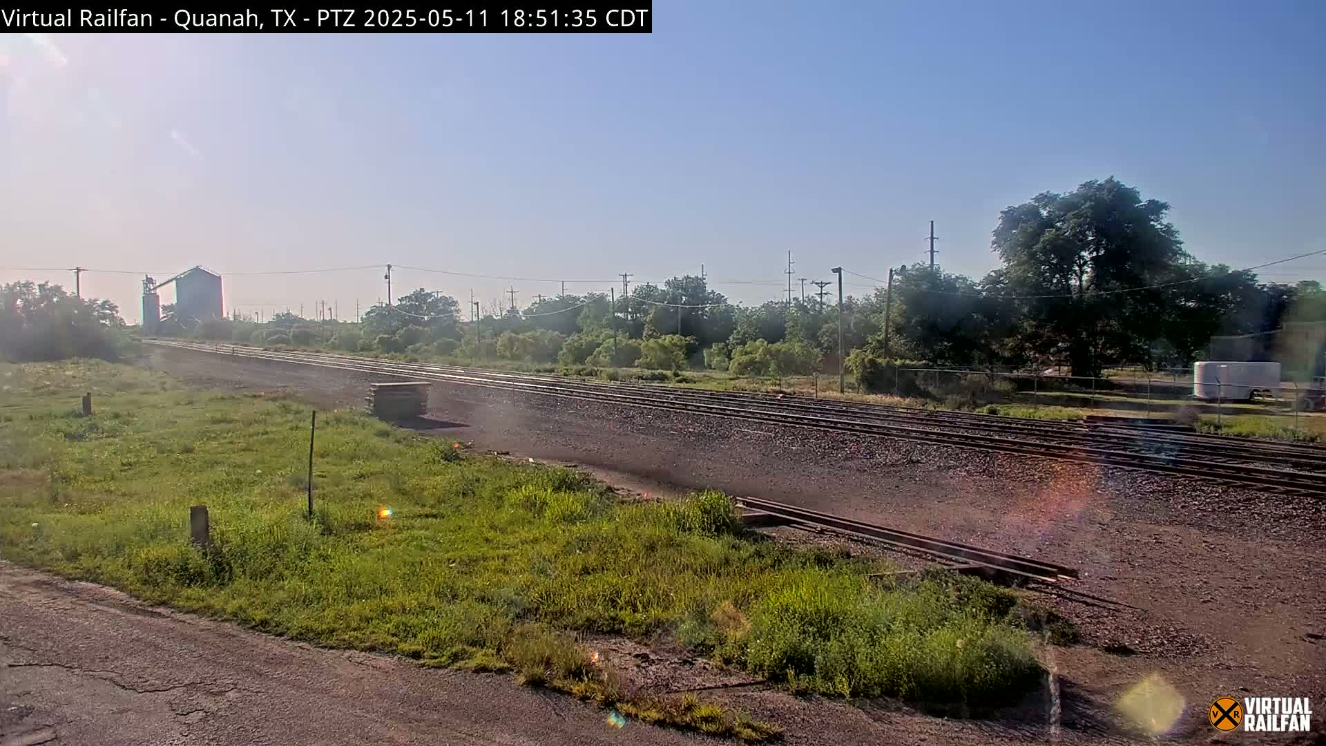 A stretch of railway tracks under a clear, bright sky passes through a grassy area with some trees and a building in the distance.