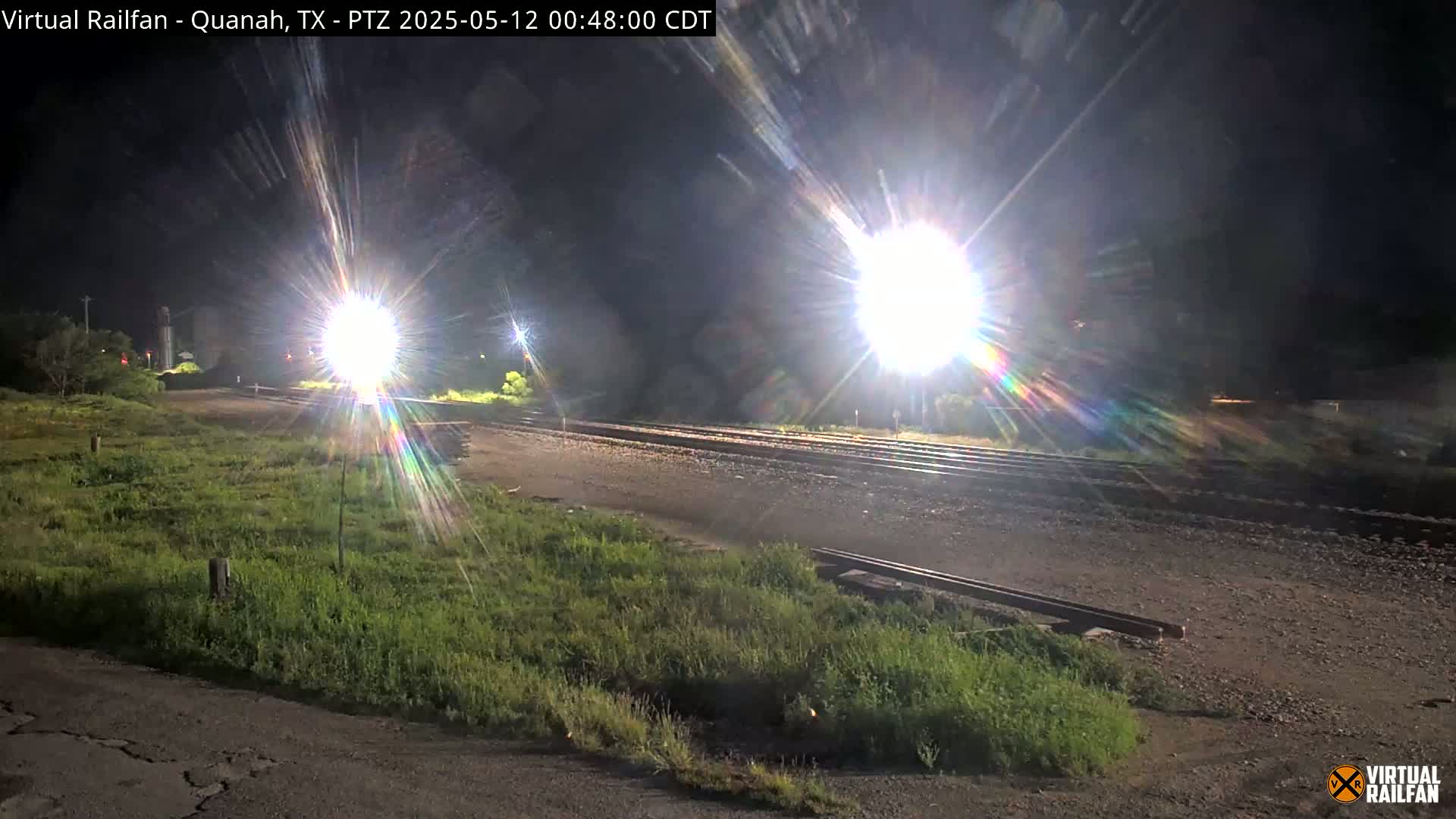 A nighttime view of railroad tracks and surrounding grassy areas, illuminated by bright lights.