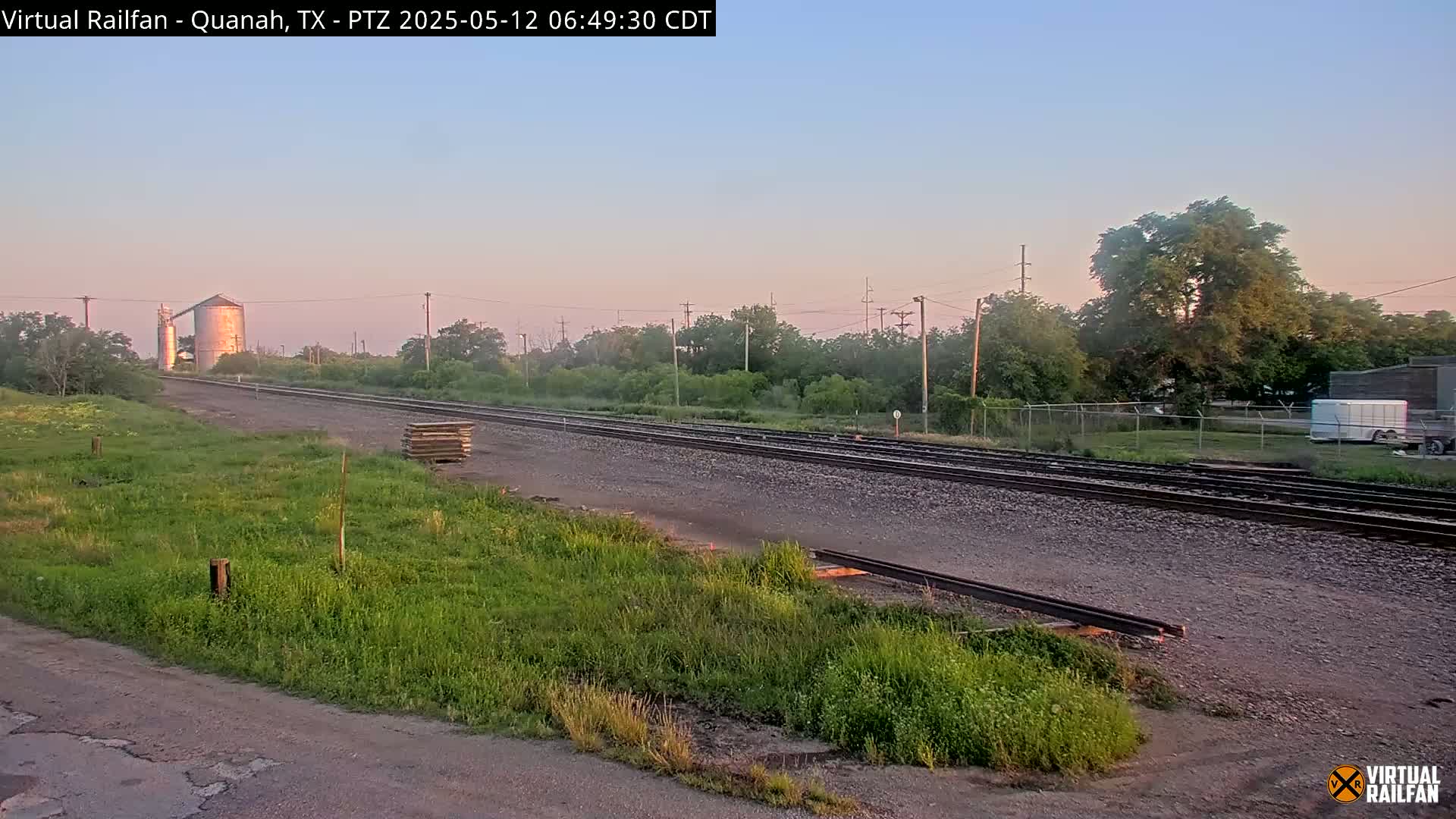 A set of train tracks stretches into the distance under a pale pink and blue sunrise sky, past a grassy field, a silo, and some trees.
