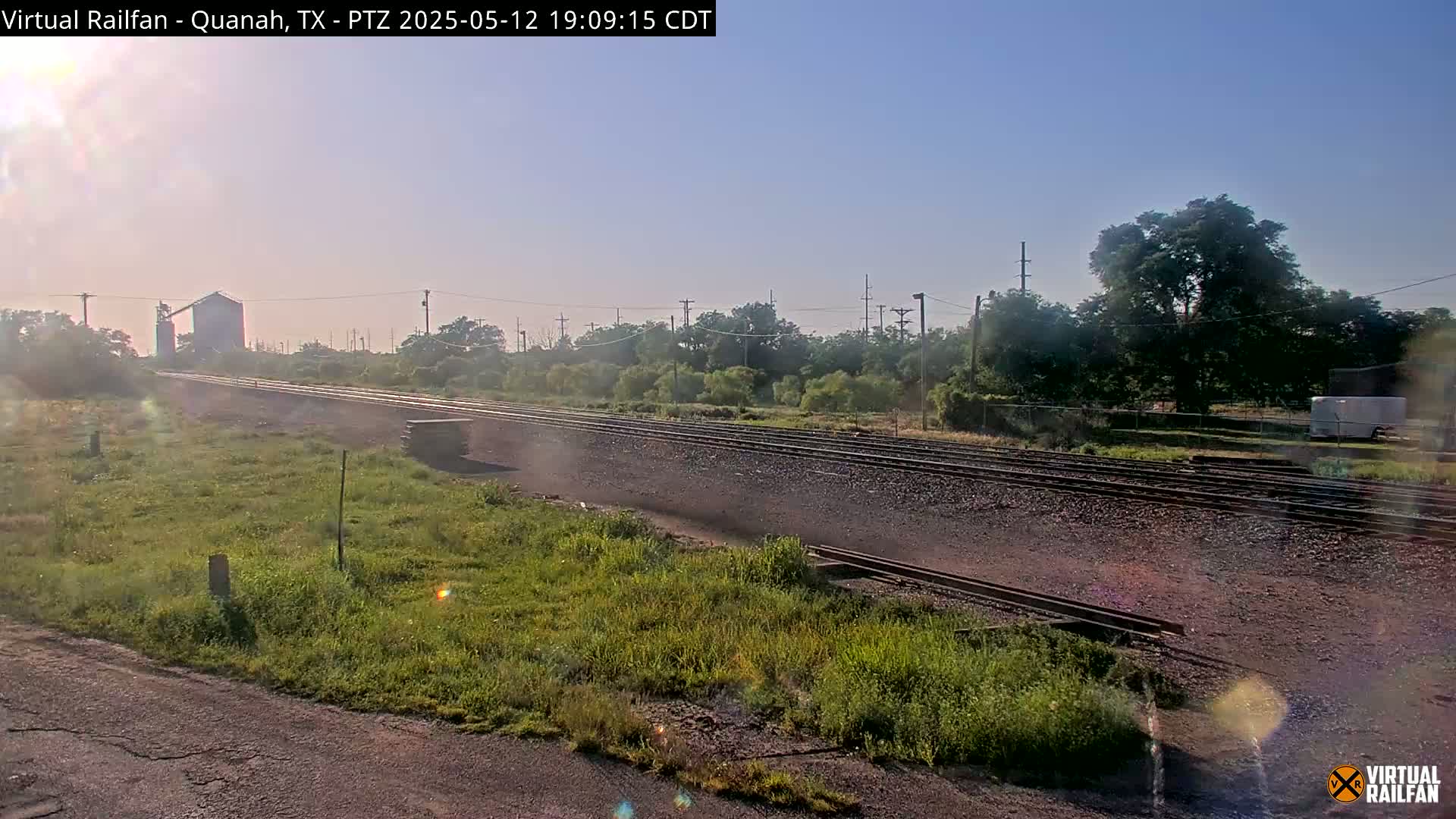 A long stretch of train tracks runs through a grassy area under a clear, bright sky, with trees and a distant building visible in the background.