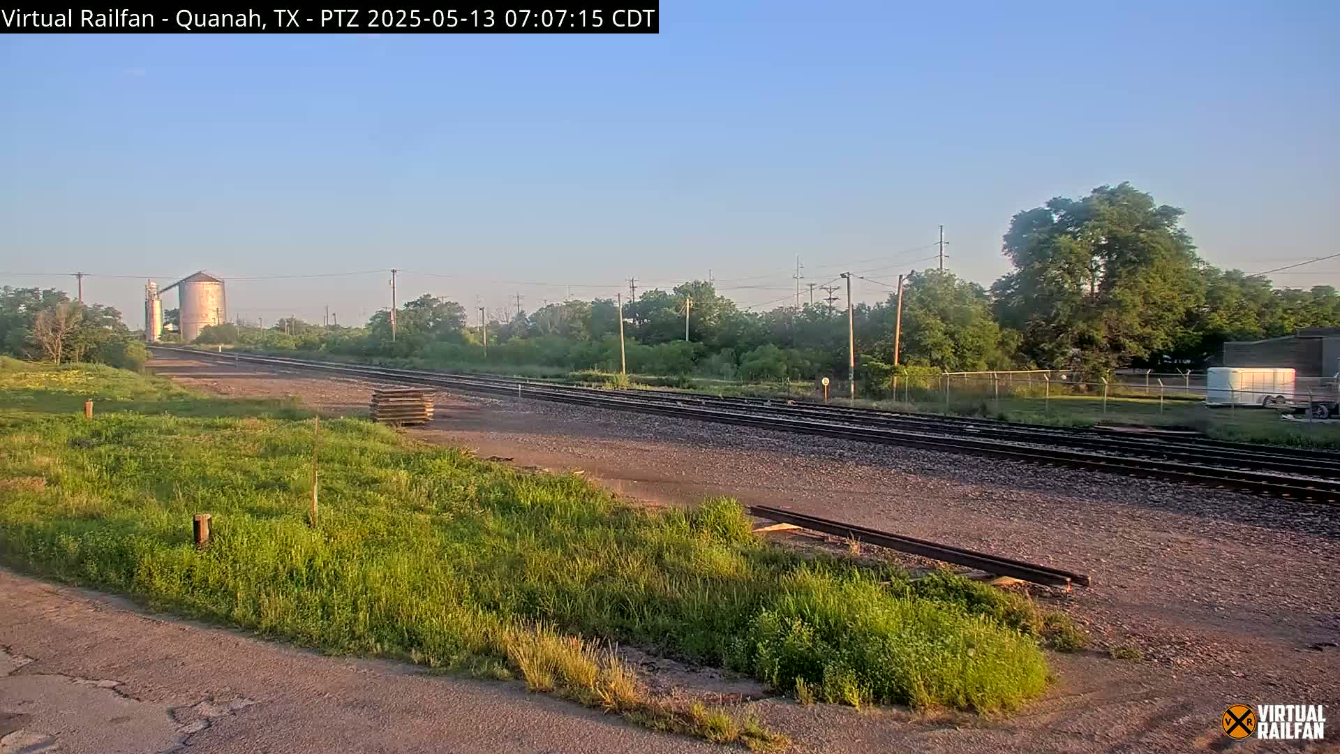 A clear, sunny day reveals a railway track stretching towards a grain silo, flanked by grassy fields and trees.