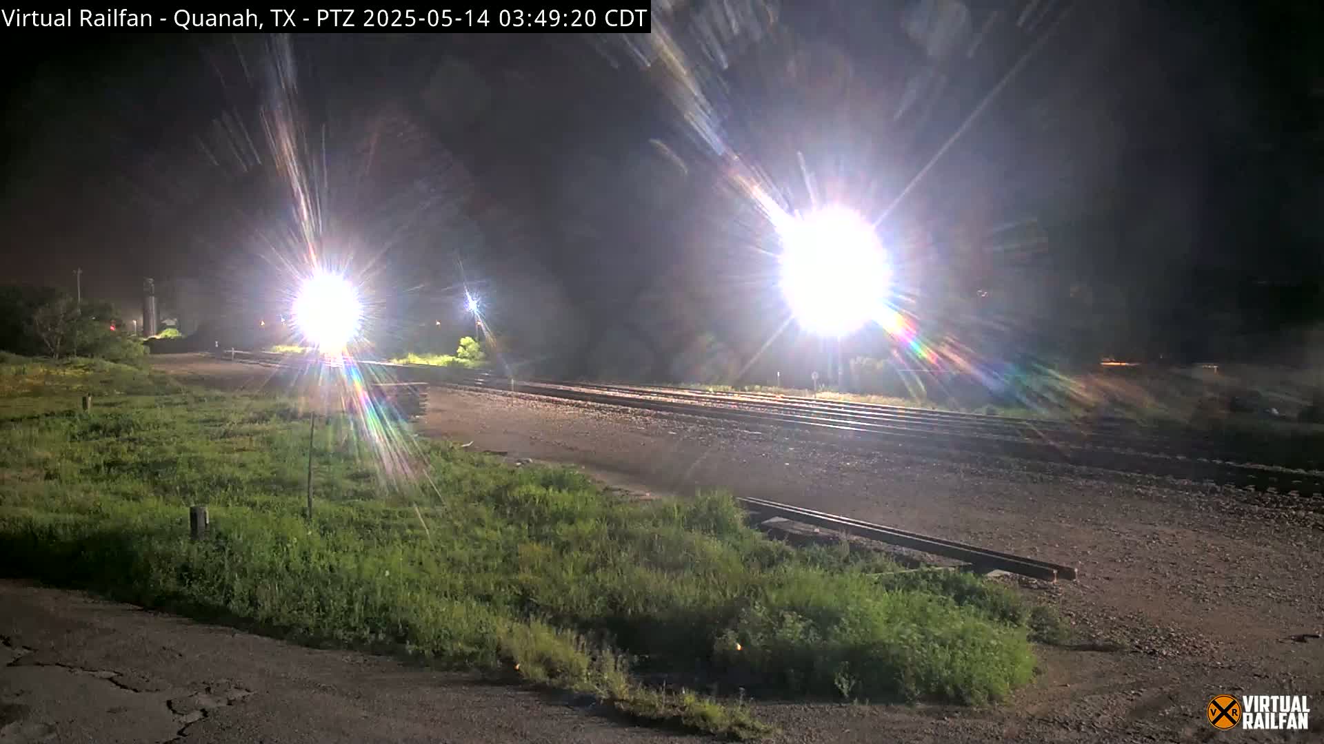 At night, bright lights illuminate railroad tracks and surrounding vegetation.