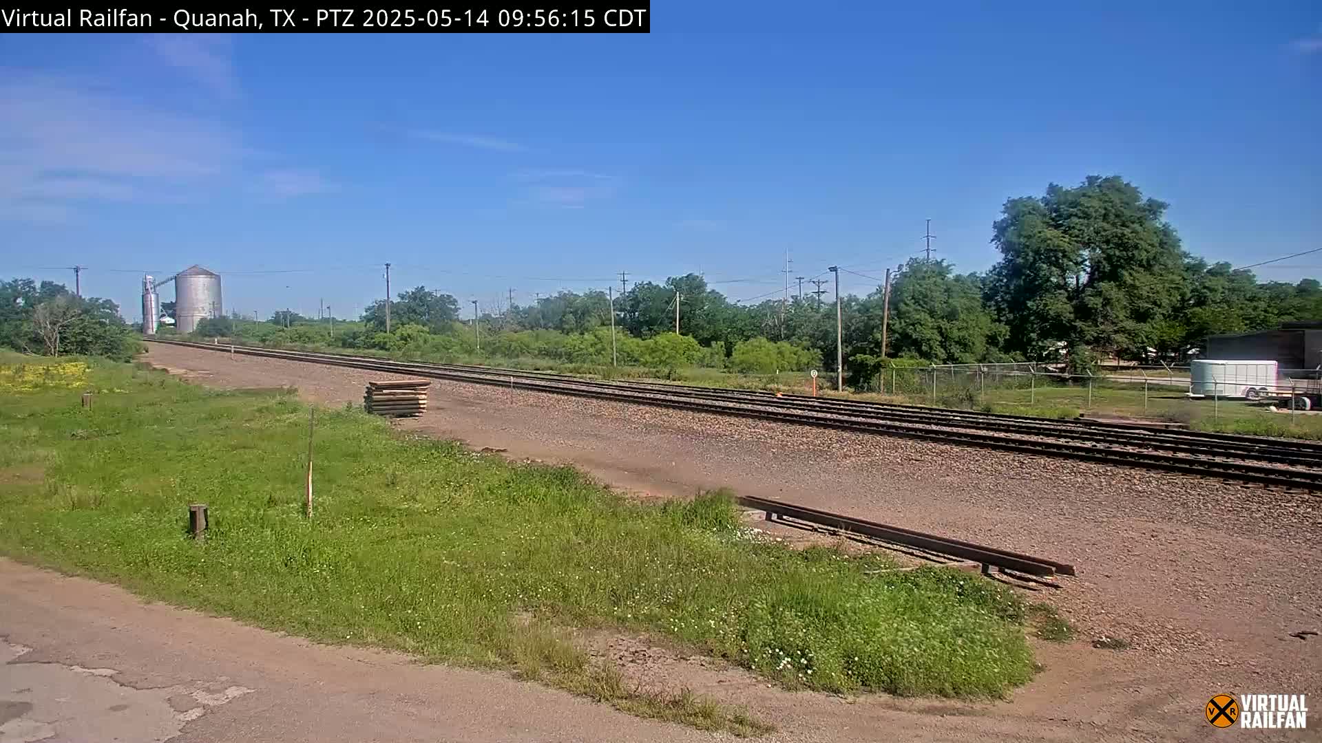 Under a clear blue sky, a railroad track stretches toward a grain silo in a grassy, rural landscape.
