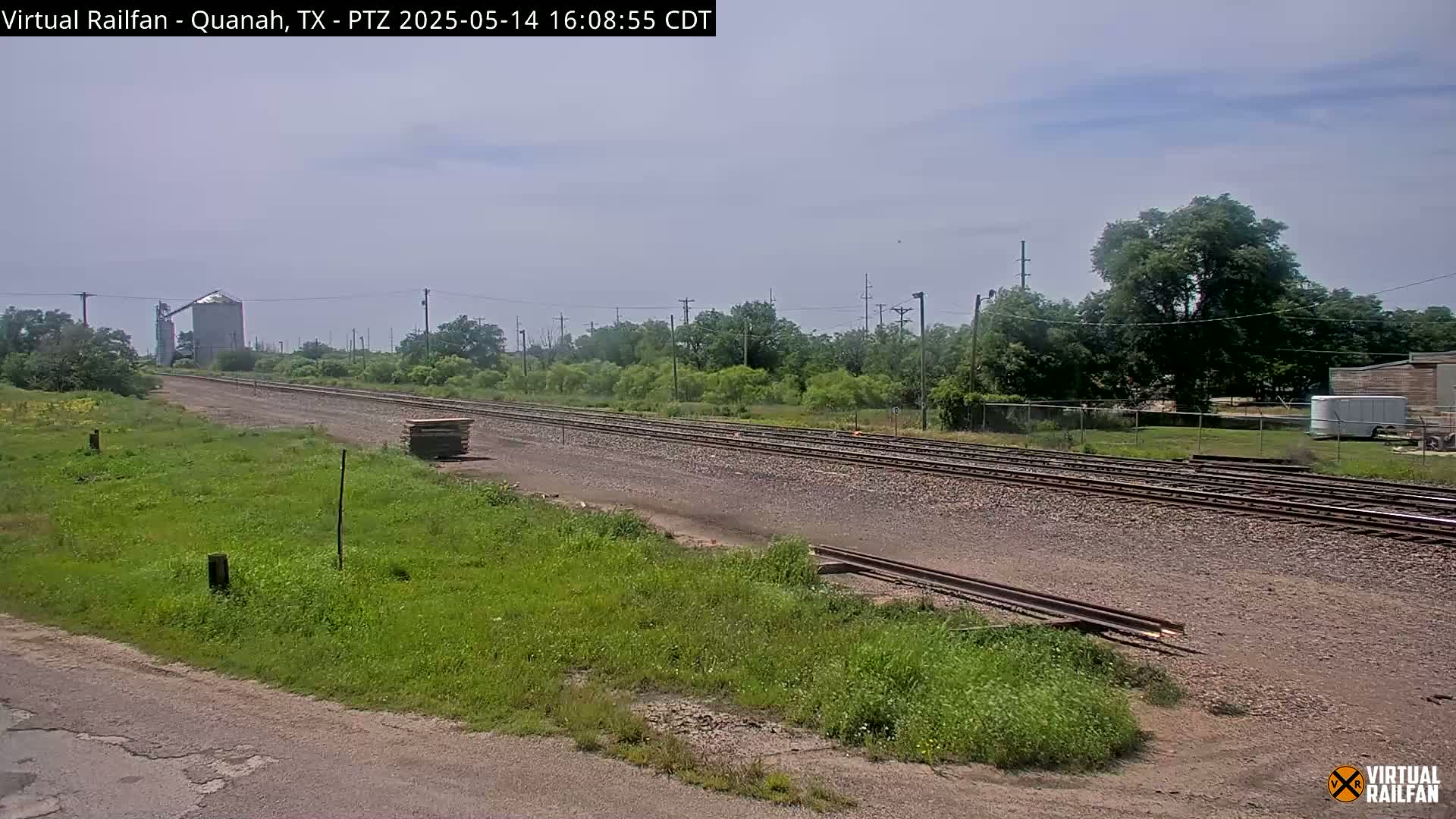A long stretch of railway tracks runs through a grassy area, under a mostly clear sky, past a grain silo and some trees.