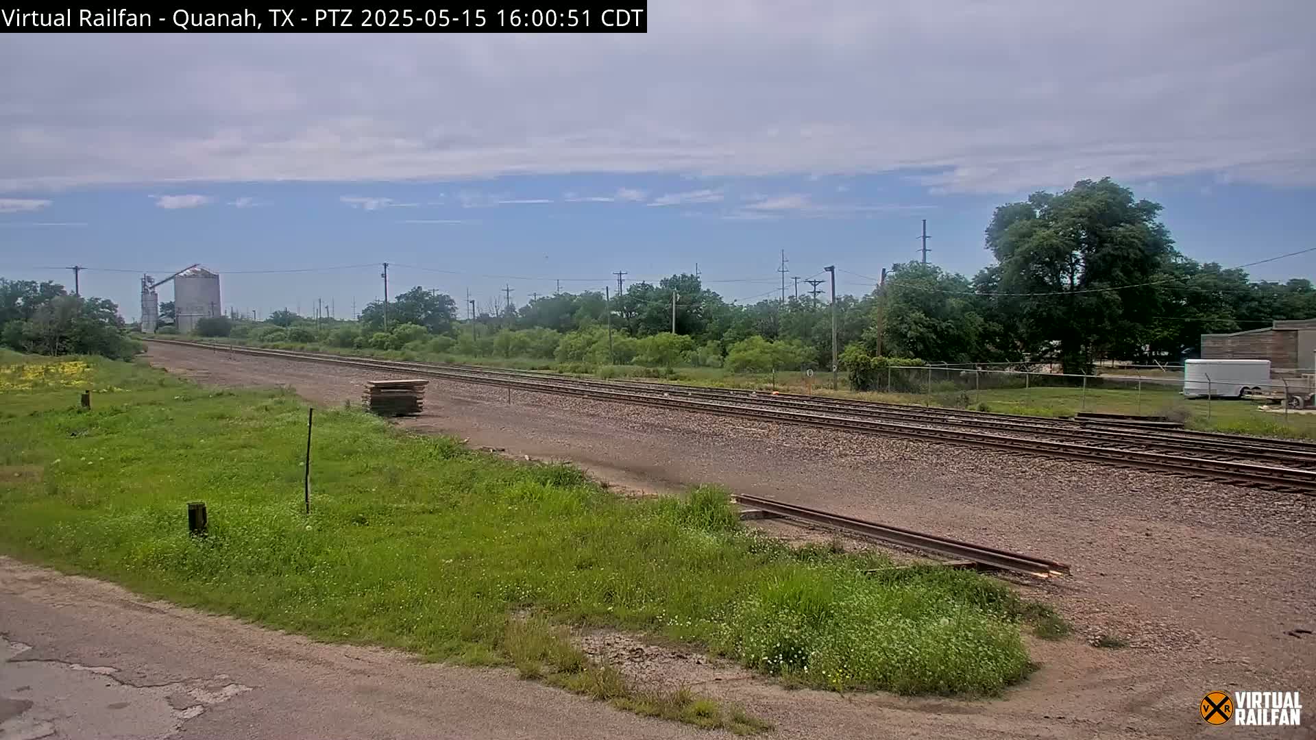 A mostly sunny day shows a view of railroad tracks extending into the distance, past green vegetation and a large silo, with a few buildings visible in the background.