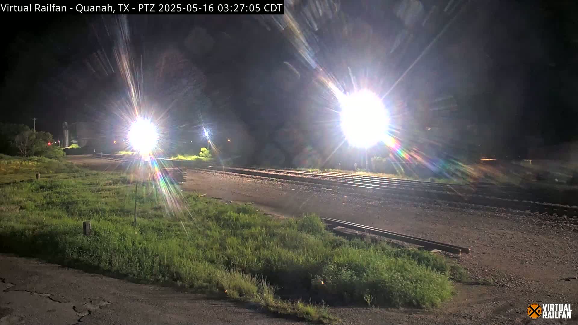 At night, bright lights illuminate railroad tracks and surrounding grassy areas.