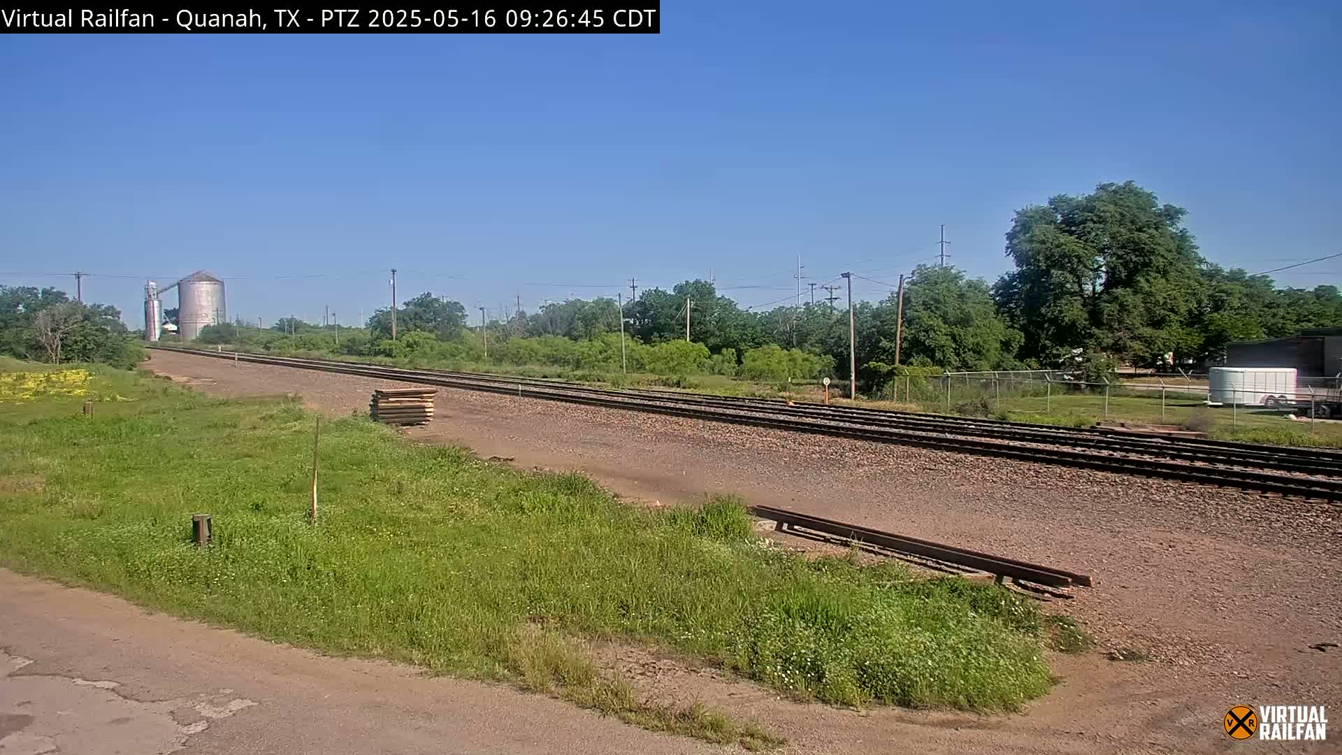 A long stretch of train tracks extends into the distance under a clear blue sky, flanked by grassy areas, trees, and a grain silo in the background.