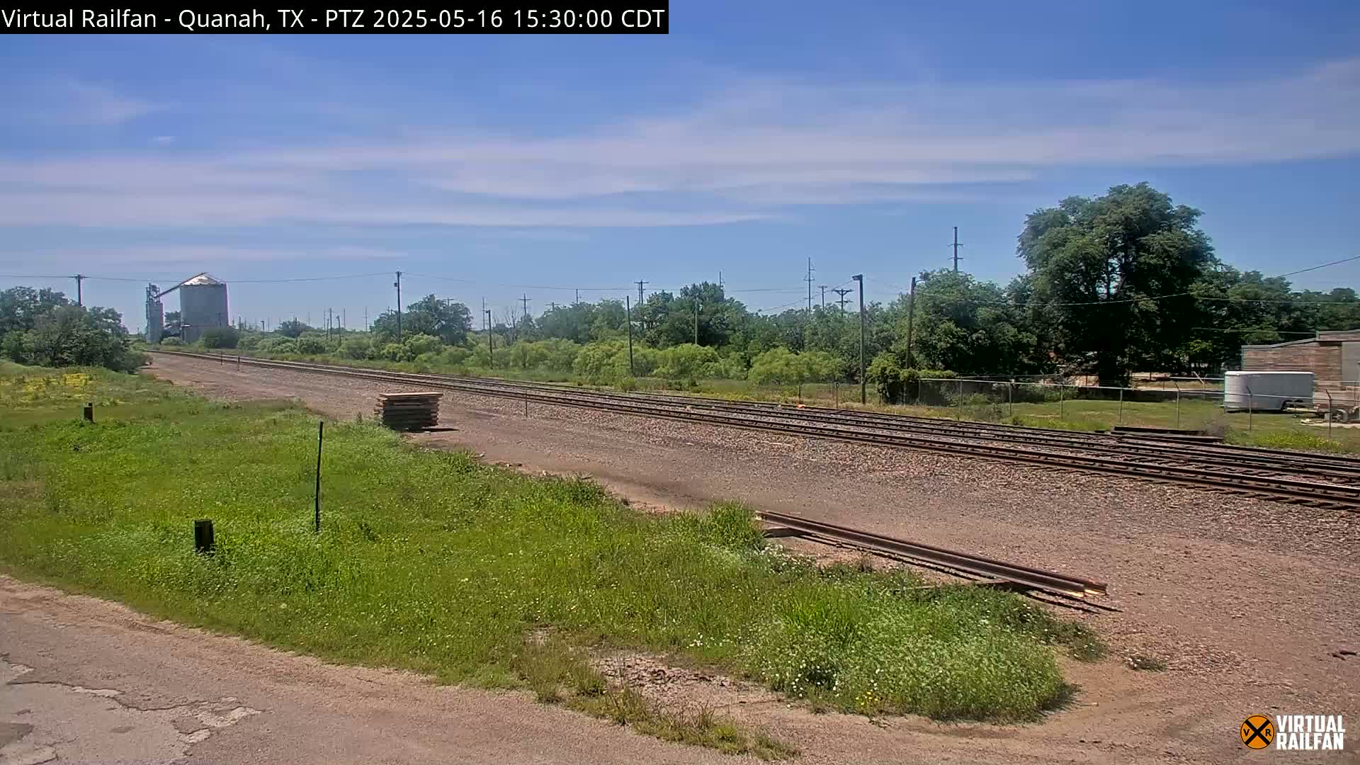 A mostly sunny day shows a view of railroad tracks extending into the distance, flanked by green vegetation, with a grain silo visible in the background.